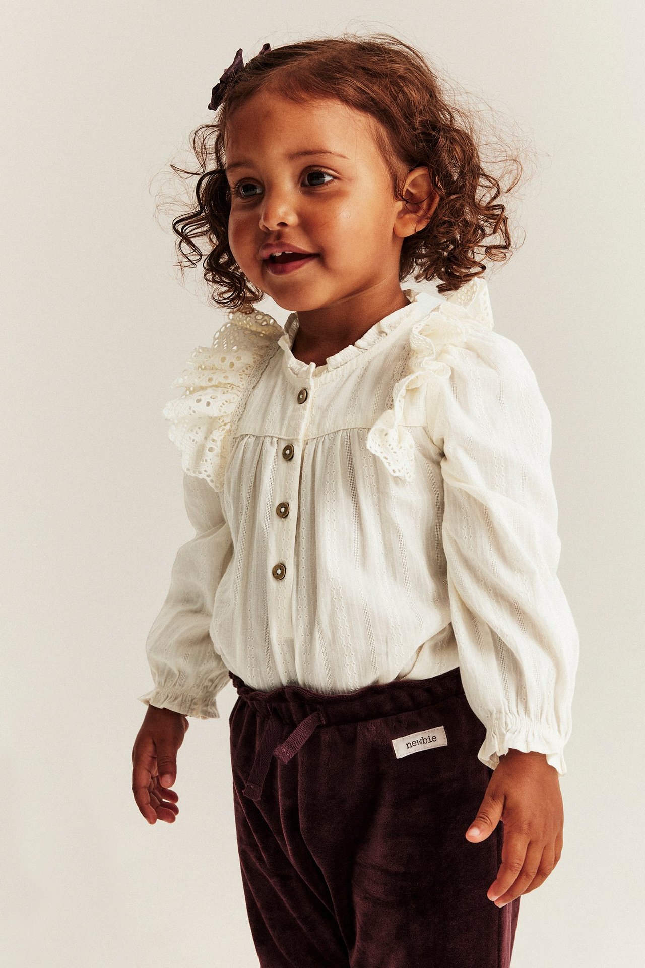 Widok z przodu: dziecko w ecru bawełnianym body z haftowanymi falbankami i ciemnofioletowych welurowych spodniach.