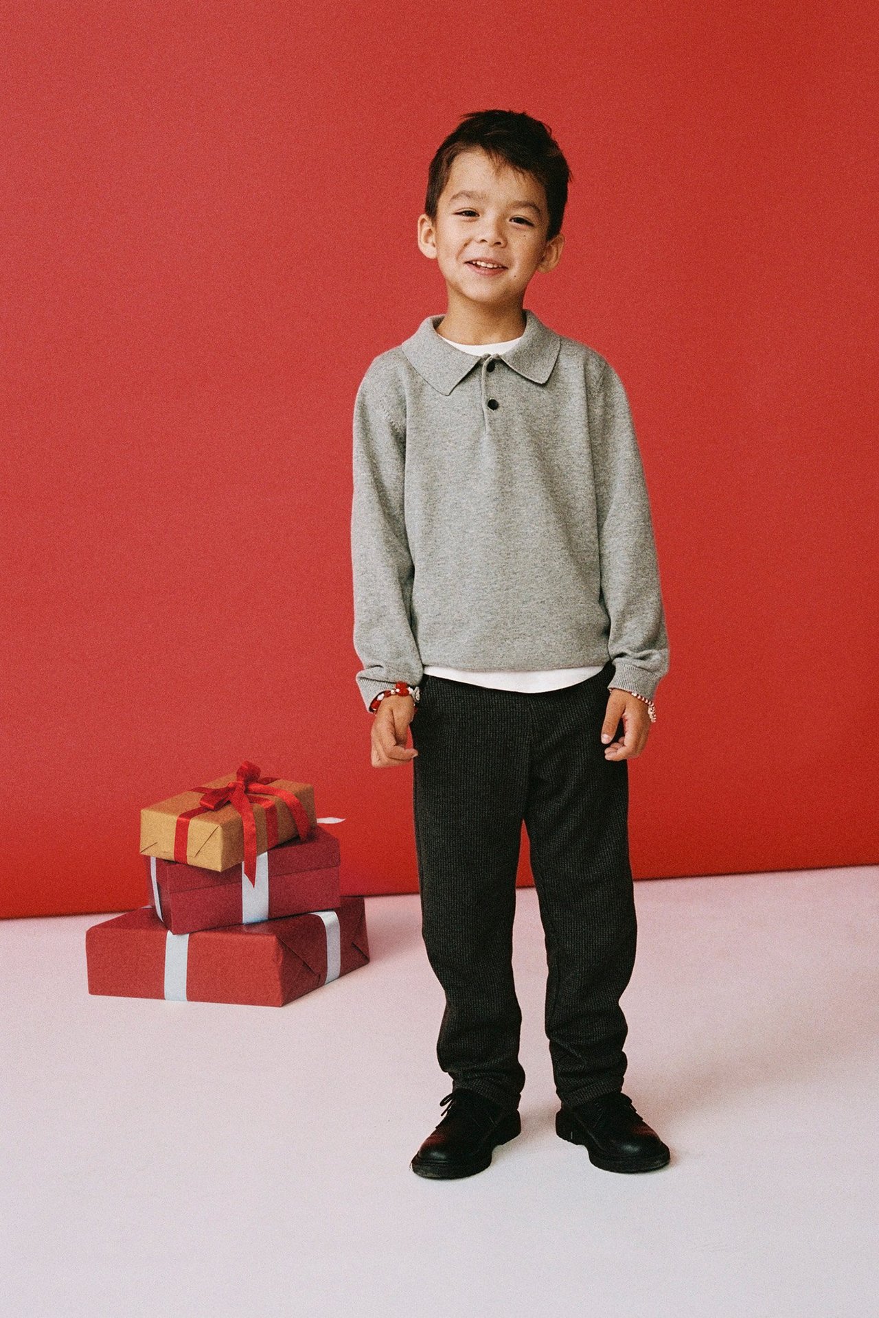 Framsida på leende barn i mörkgrå finstickad pikétröja av bomull, mörkgrå byxor och svarta skor.