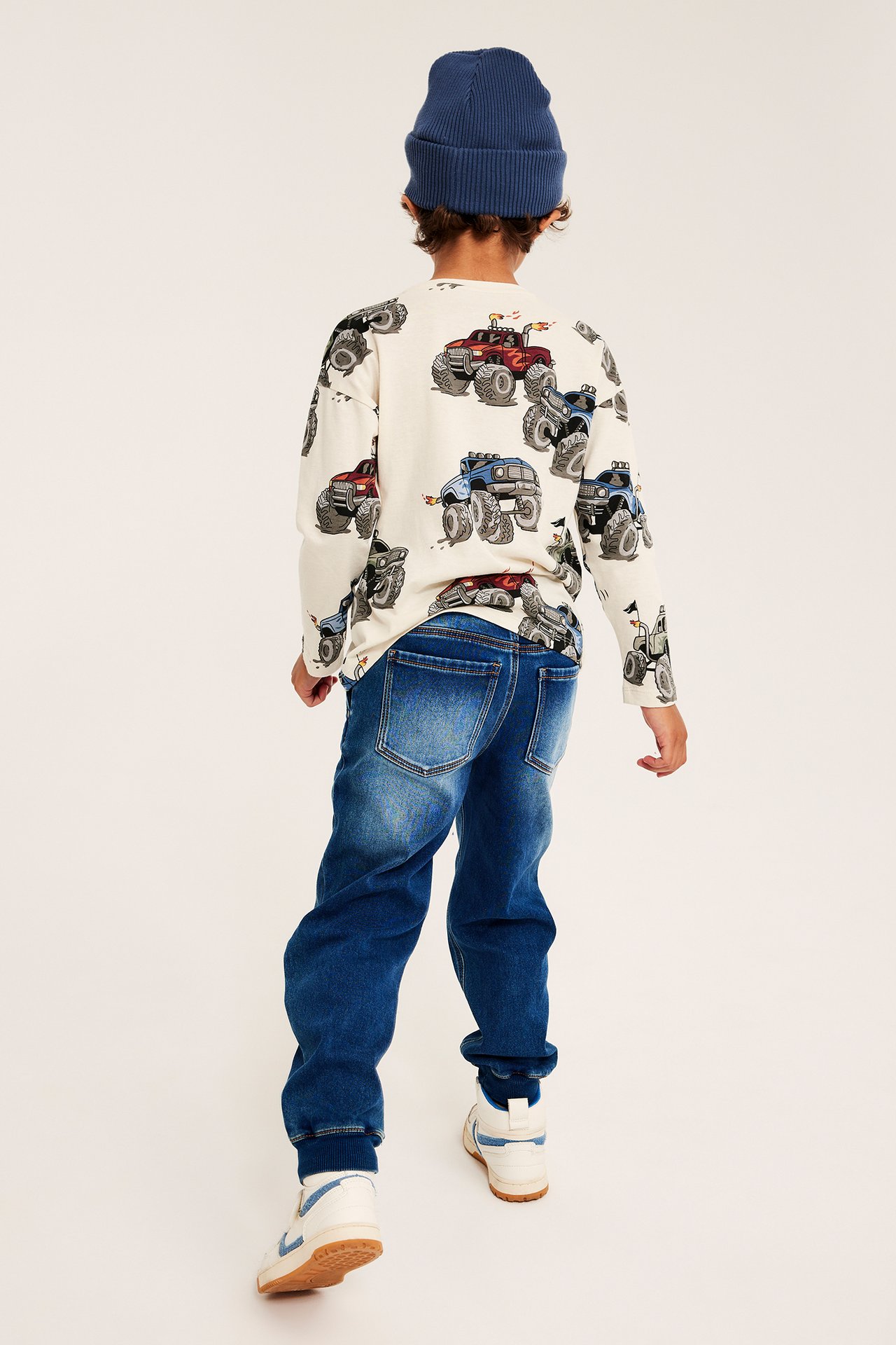 Ryggvy av ett barn i benvit långärmad t-shirt med monstertrucktryck, blå mössa, jeans och sneakers.