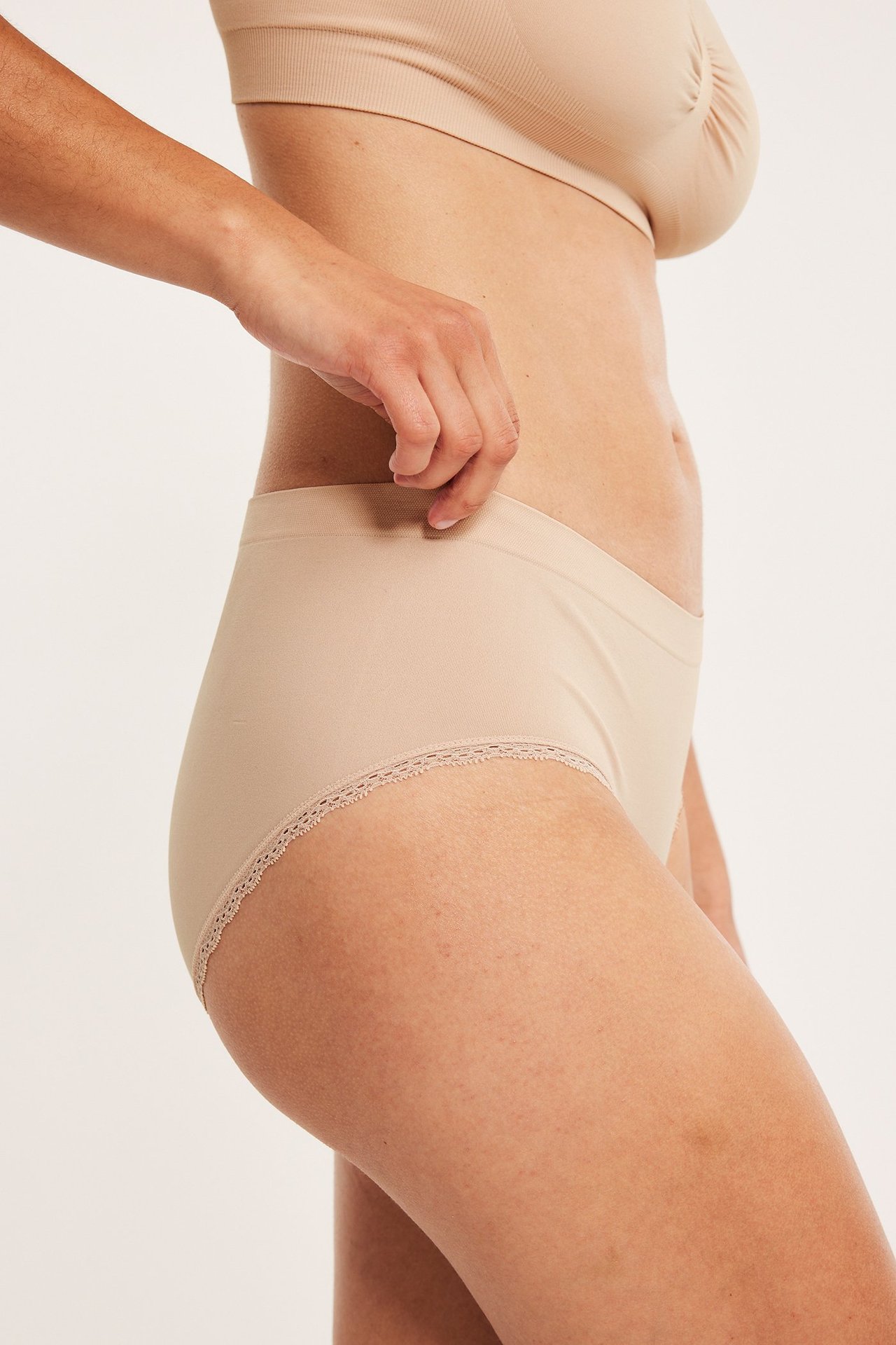 Sidovy av kvinna i ljusbeige sömlös hipstertrosa med spetskant och matchande sömlös BH.