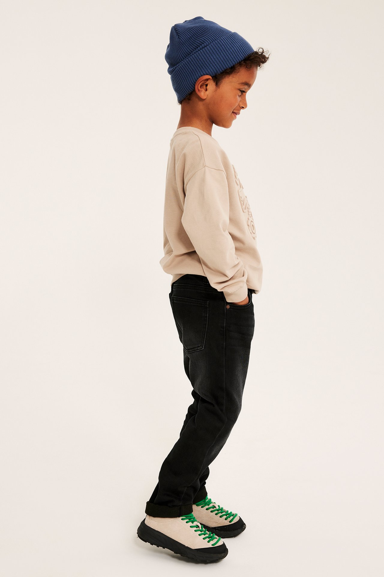 Barn med blå mössa, beige sweatshirt, svarta jeans och beiga skor med gröna skosnören, sidovy.