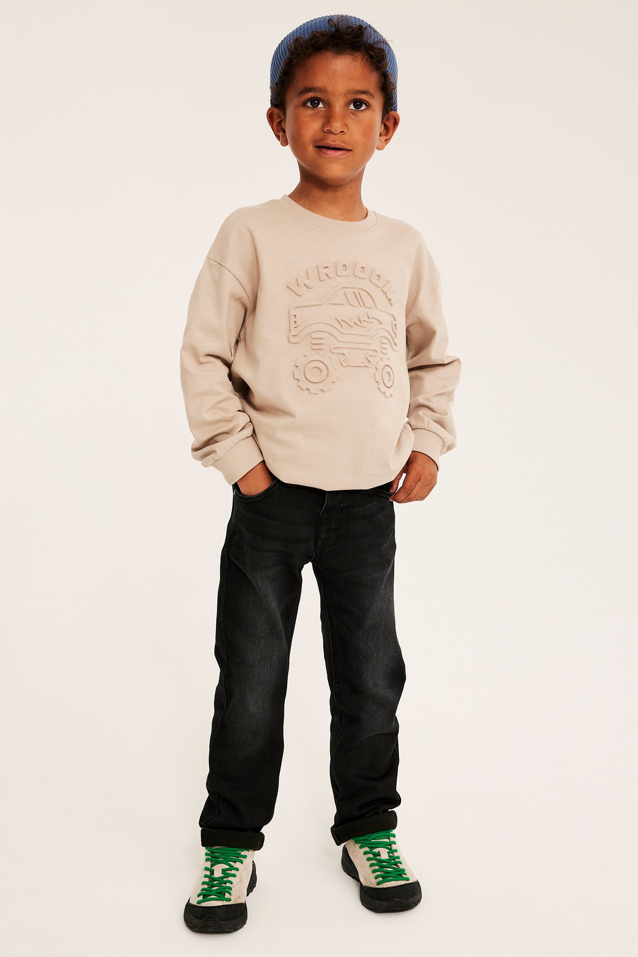 Ljusbrun präglad monstertrucktröja, mörka jeans, blå mössa och sneakers på pojke.