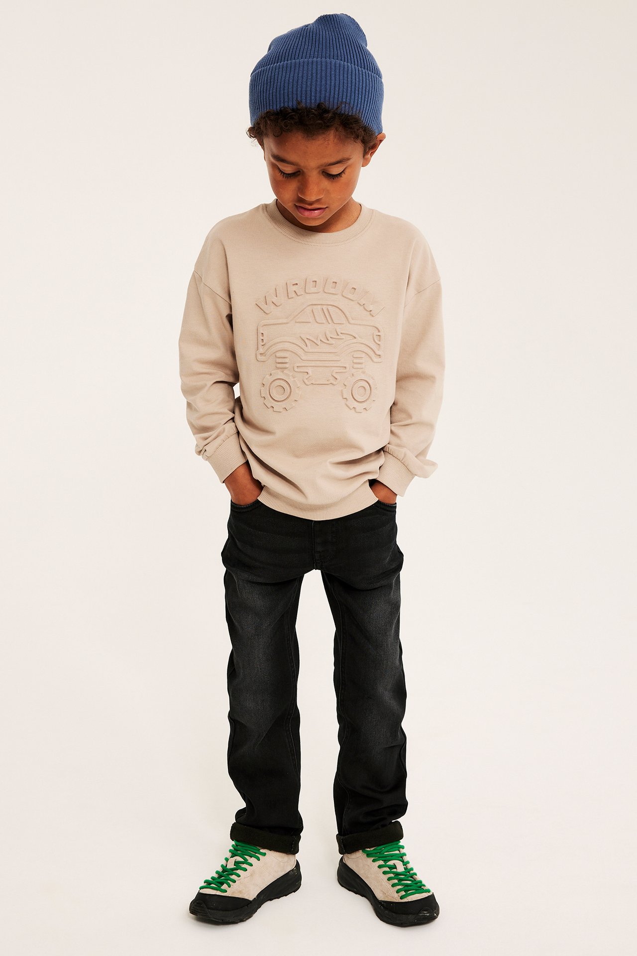 Framsida: Barn i ljusbeige präglad monstertrucktröja, blå mössa, svarta jeans, gröna sneakers.