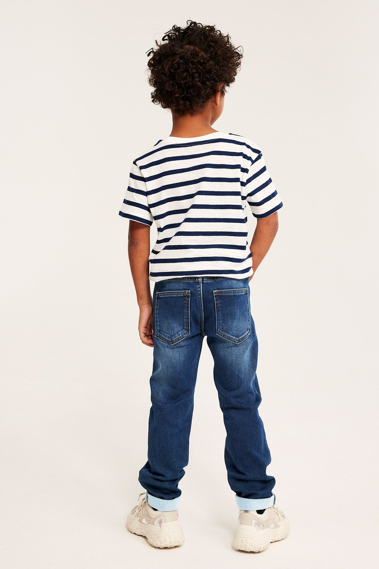Ryggvy av ett barn i mörkblå raka joggerjeans, en randig t-shirt och beigefärgade sneakers.