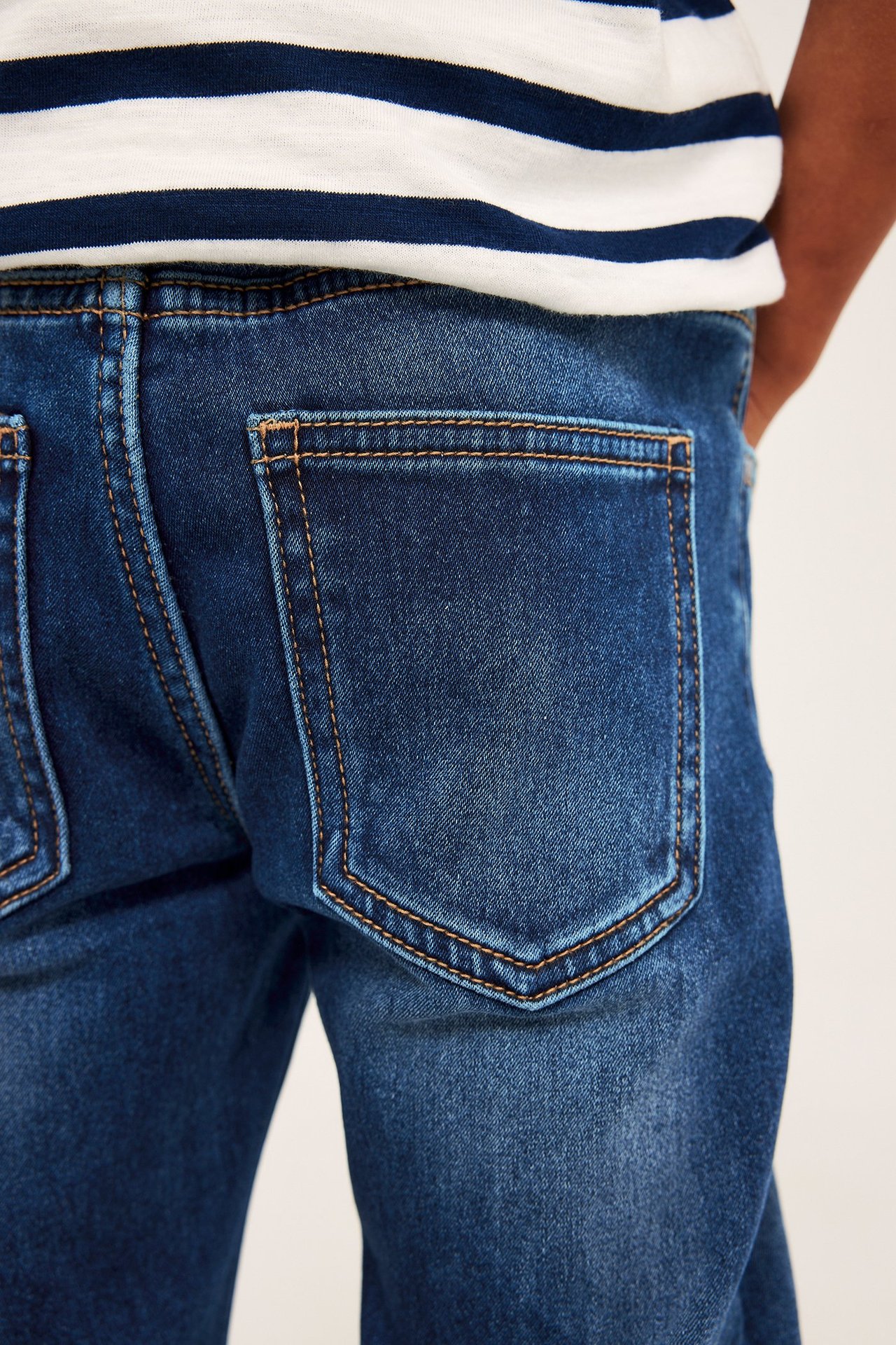 Bakifrån av ett barn i mörkblå raka joggerdenimjeans med orange sömmar och en randig t-shirt.