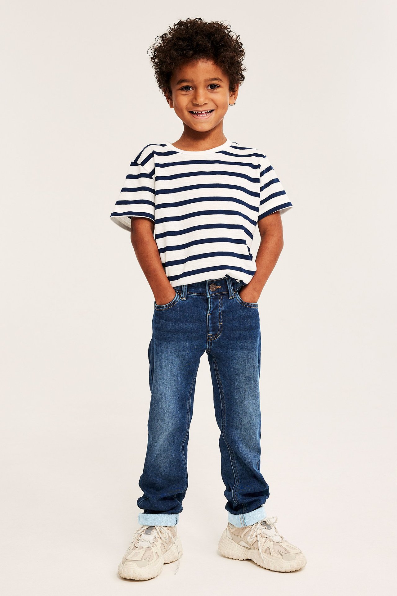 Frontbilde av et barn i mørkeblå rette jogger-denimjeans, en stripete t-skjorte og beige joggesko.
