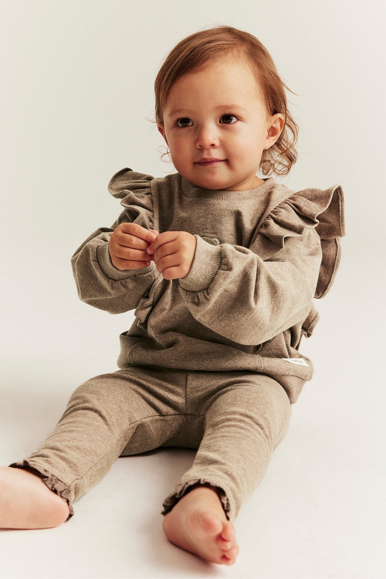 Forfra: Baby i brune melange leggings med blondekant og matchende langermet topp med volanger.