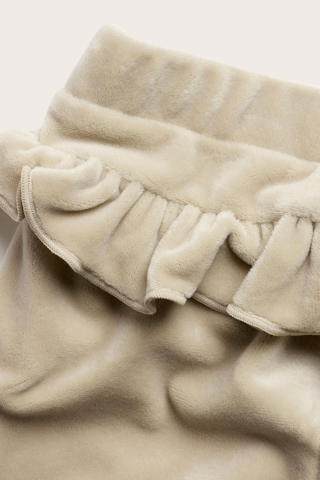 Nærbilde av beige velour babyjoggebukser med volangdetalj.