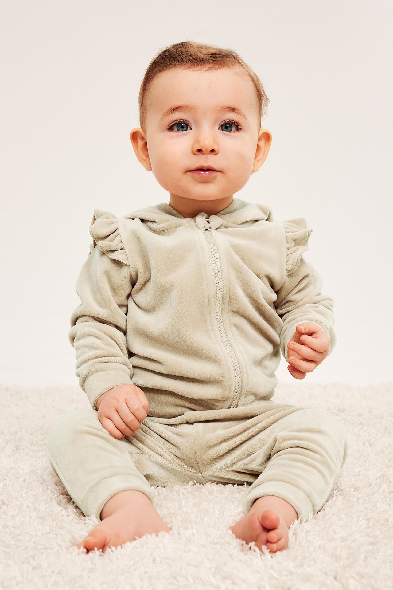 Forfra, baby i beige velour-joggedress med glidelåshettegenser og volanger på skuldrene.
