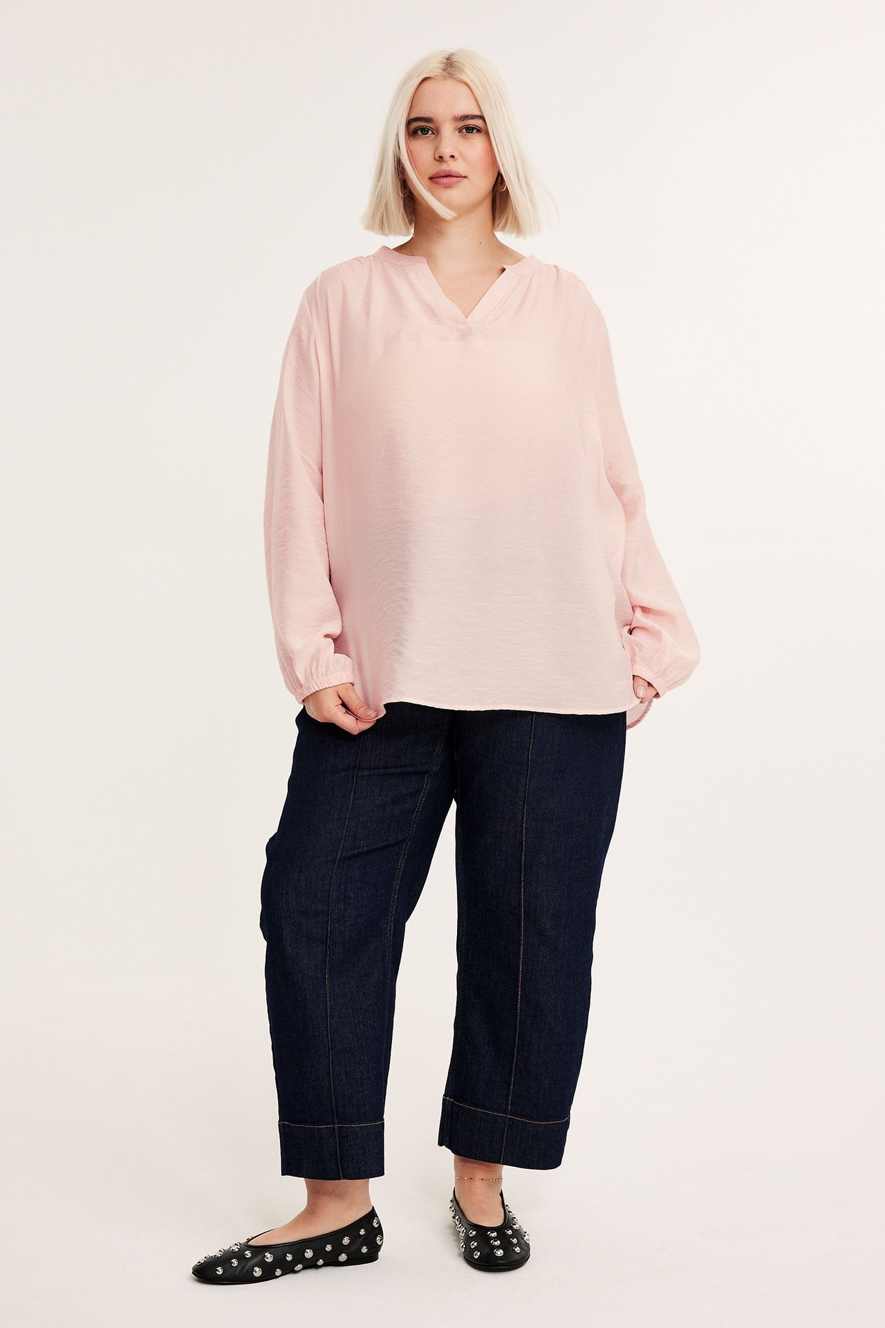 Edestä: nainen yllään vaaleanpunainen strukturoitu pusero, tummansiniset capri-farkut ja mustat niittikoristeiset ballerinat.