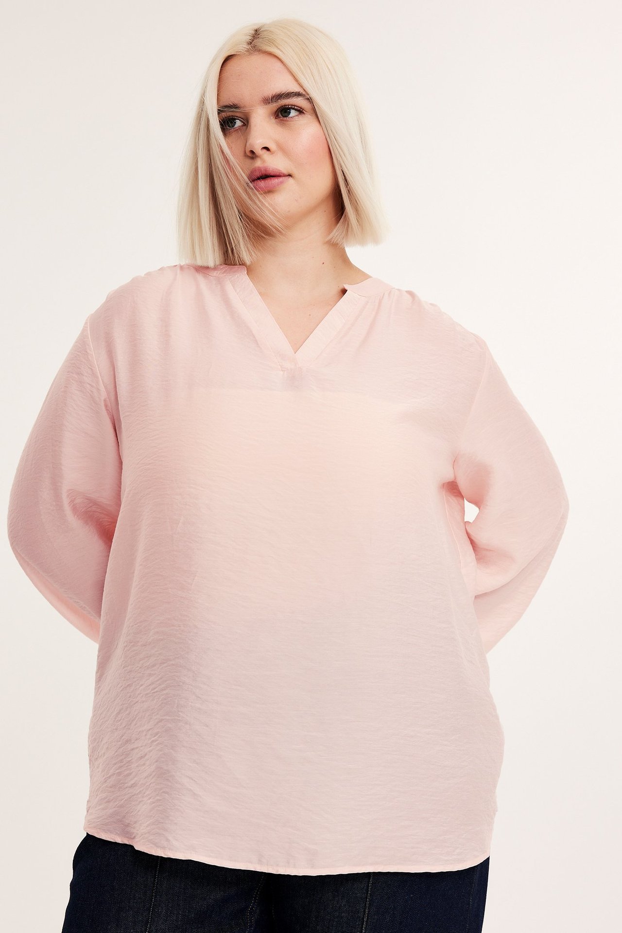 Widok z przodu: kobieta w luźnej, jasnobeżowej bluzce z dekoltem w serek i elastycznymi mankietami.