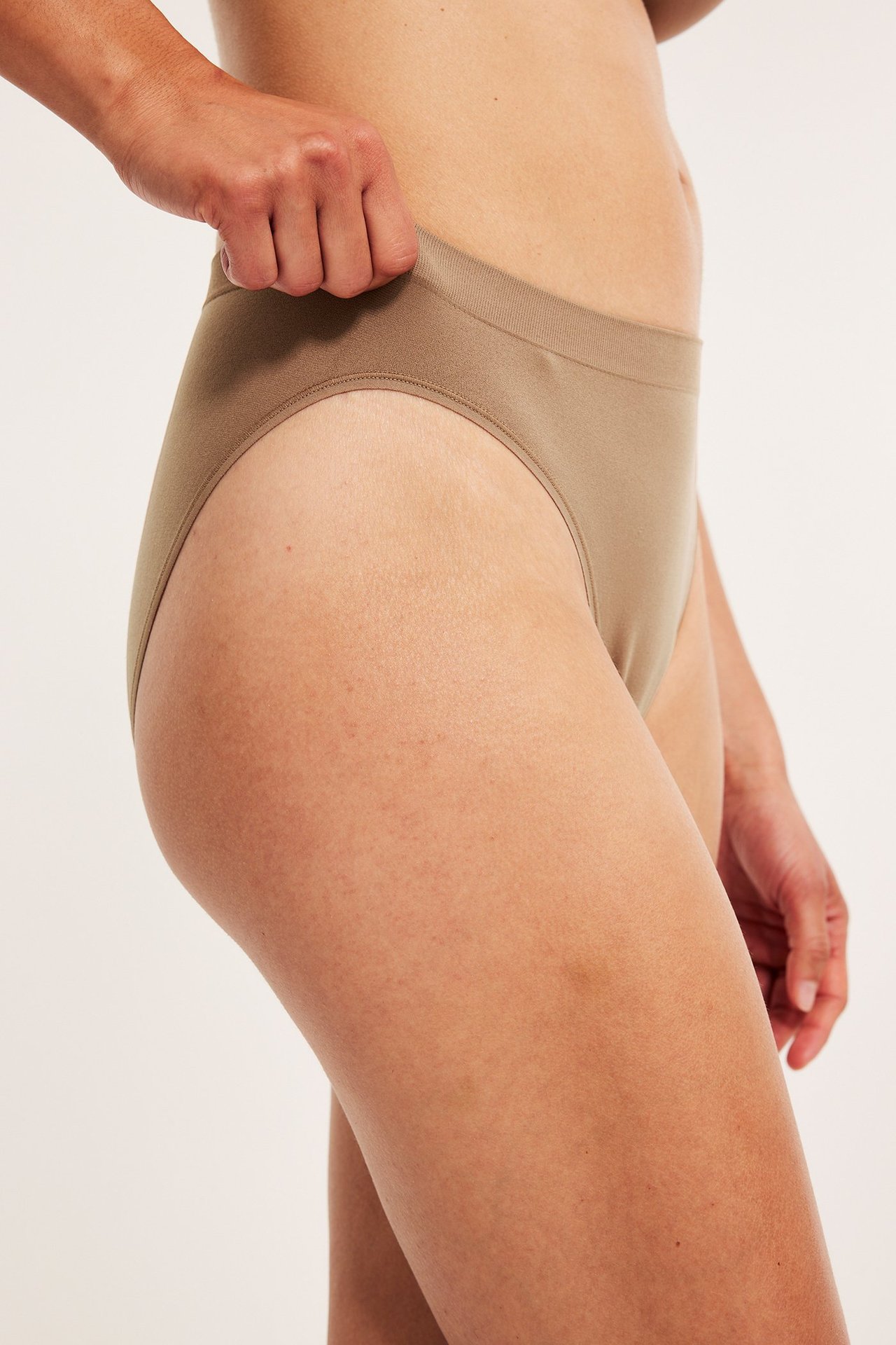 Sidebilde av en kvinne i beige truse med ribbet midje, hånden hennes trekker i midjen.