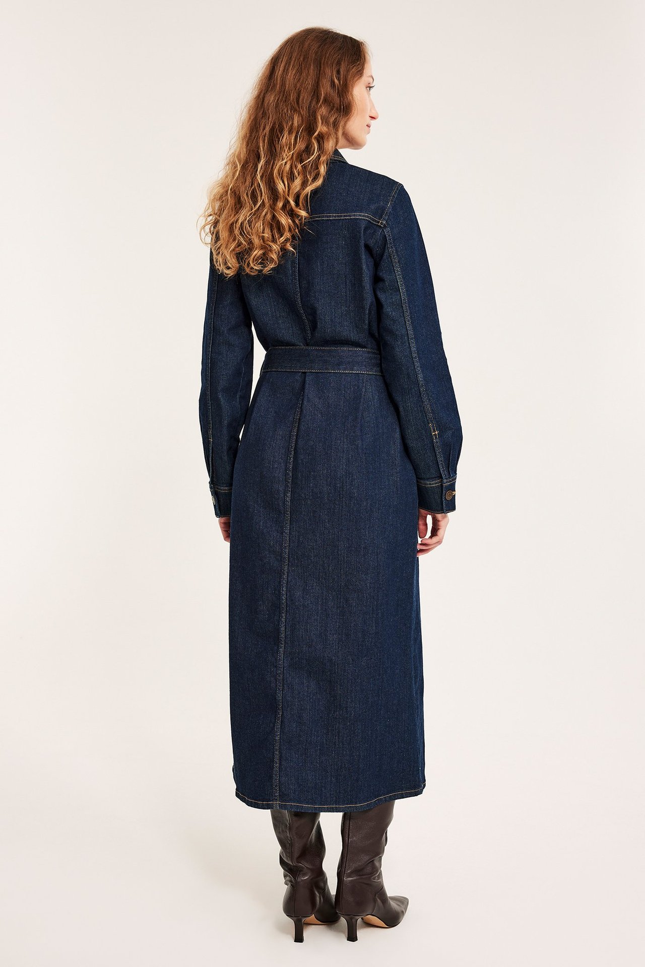 Naisen selkäpuolelta: pitkä, vyöllinen tummansininen farkkupaitamekko ja tummanruskeat korkeakorkoiset saappaat.