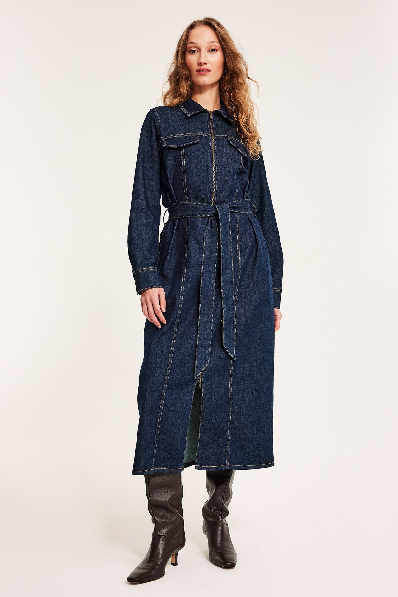 Edestä: nainen tummassa denim-midimekossa, vetoketju, solmittava vyö, tummanruskeat saappaat.