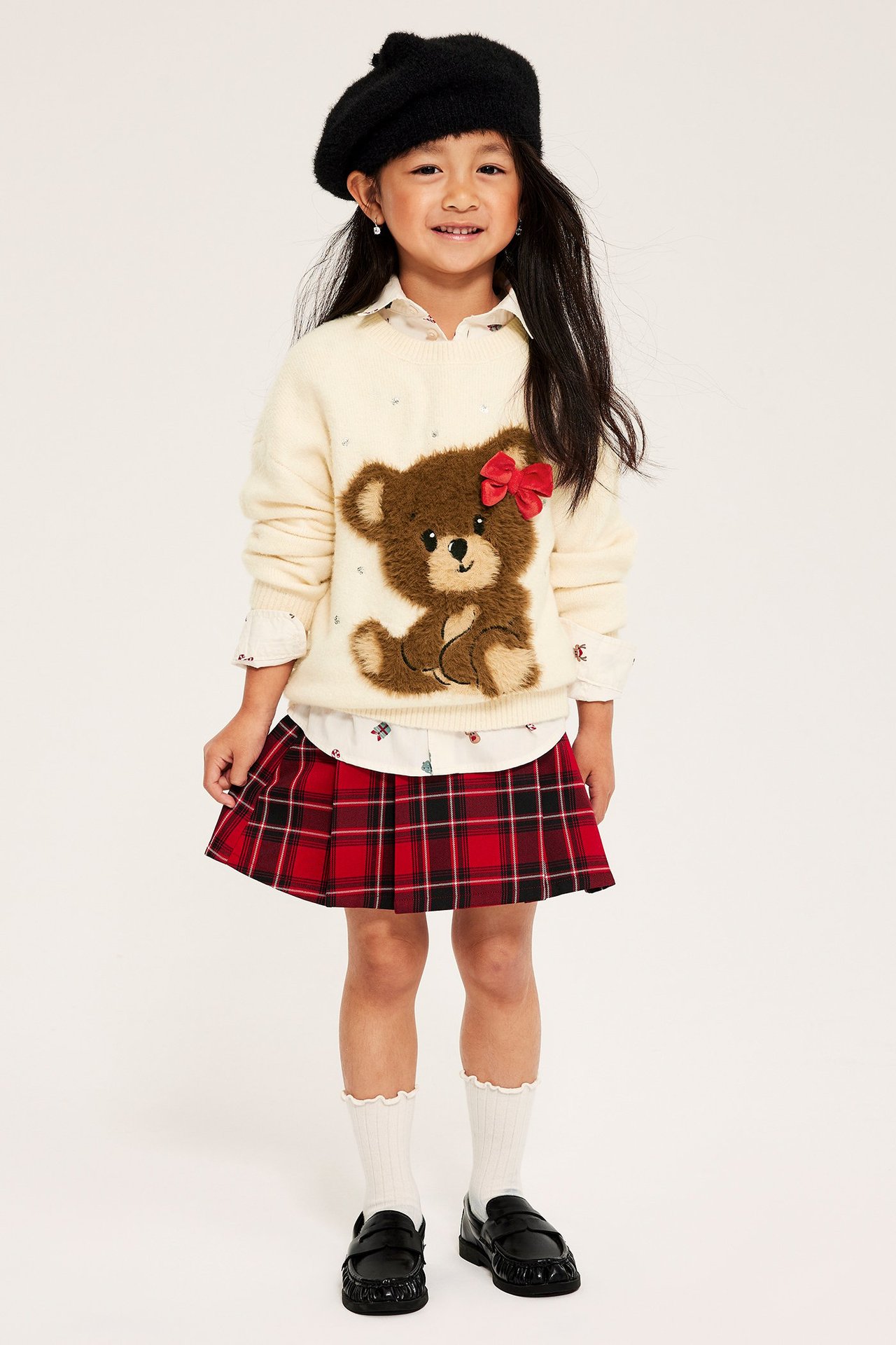 Tyttö offwhite-neuletunika, jossa applikointi ja punainen rusettin, punainen ruudullinen hame.