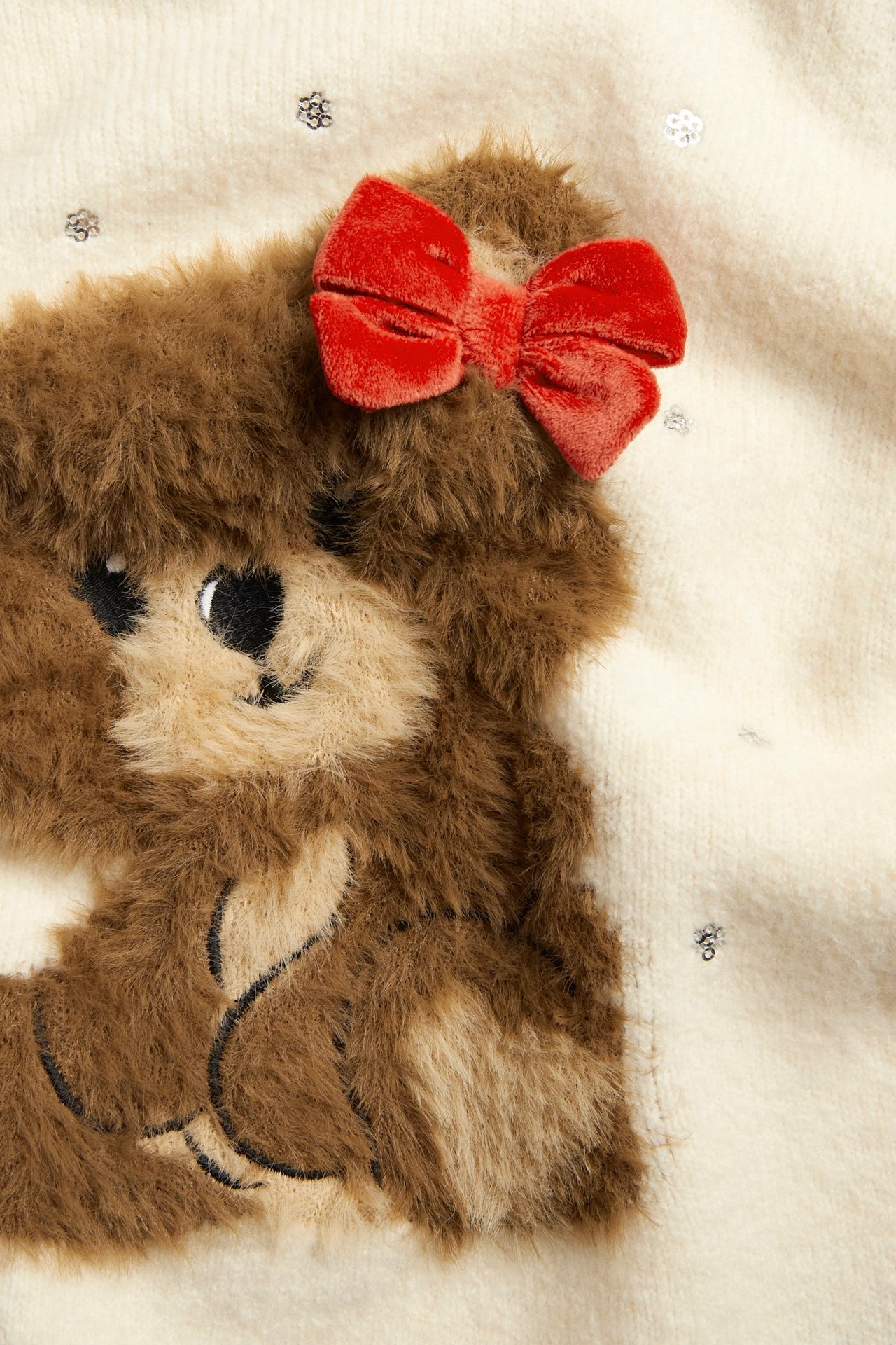 Zbliżenie: Sweter dzianinowy ecru z puszystą, brązową aplikacją misia, czerwoną kokardą i srebrnymi cekinami.