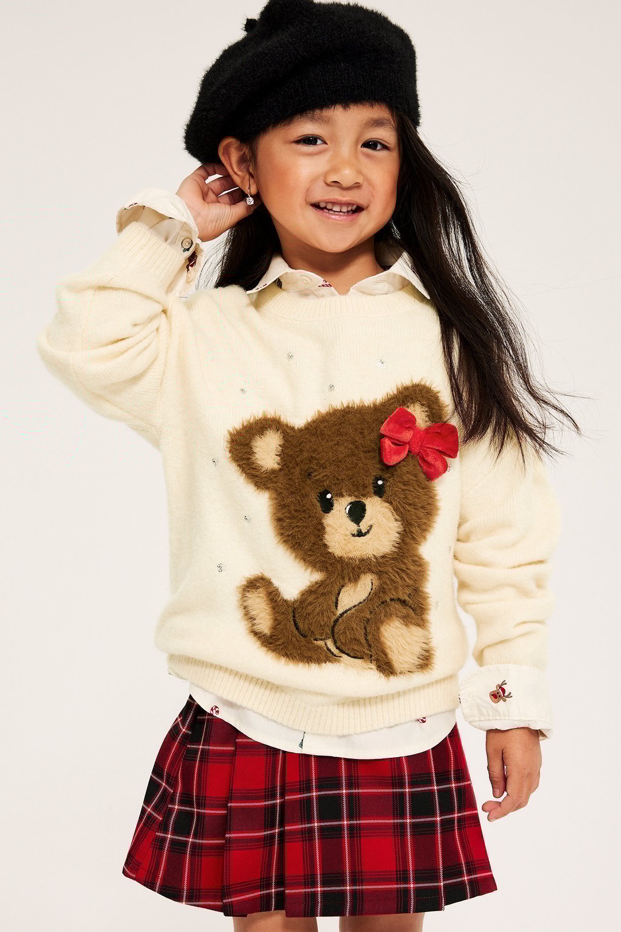 Tyttö offwhite-värinen kirjottu villapaita, punainen rusetti, punainen ruudullinen hame.