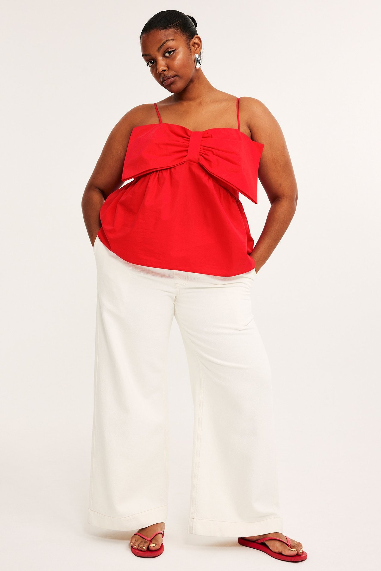 Forfra: kvinne i rød bomulls peplum-topp med sløyfe, hvite vide bukser og røde flip-flops.