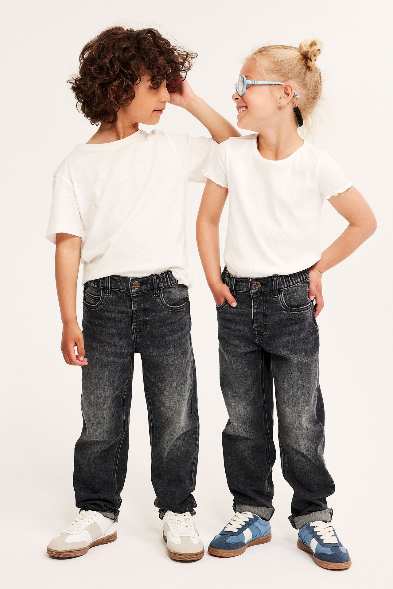 Forfra: to barn i grå relaxed-fit denimjeans, hvite t-skjorter og joggesko; jenta har briller.