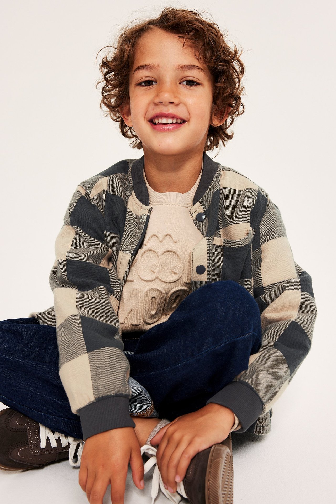 Kid bär en beige-grå rutig flanellskjorta med tryckknappar, över en beige sweatshirt.