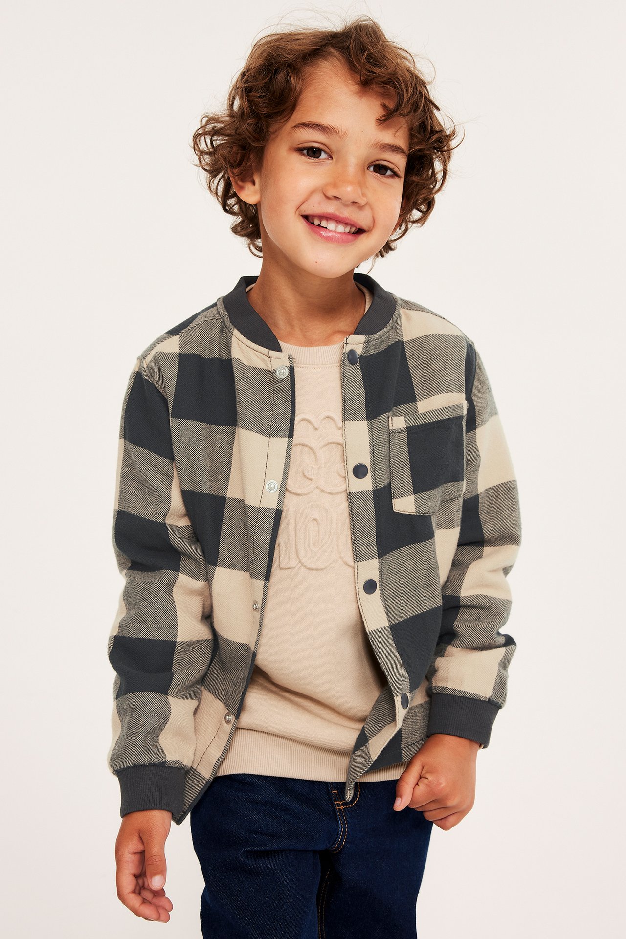 Barn har på seg en lys beige og mørkegrå rutete flanellskjorte, beige genser og mørkeblå jeans.