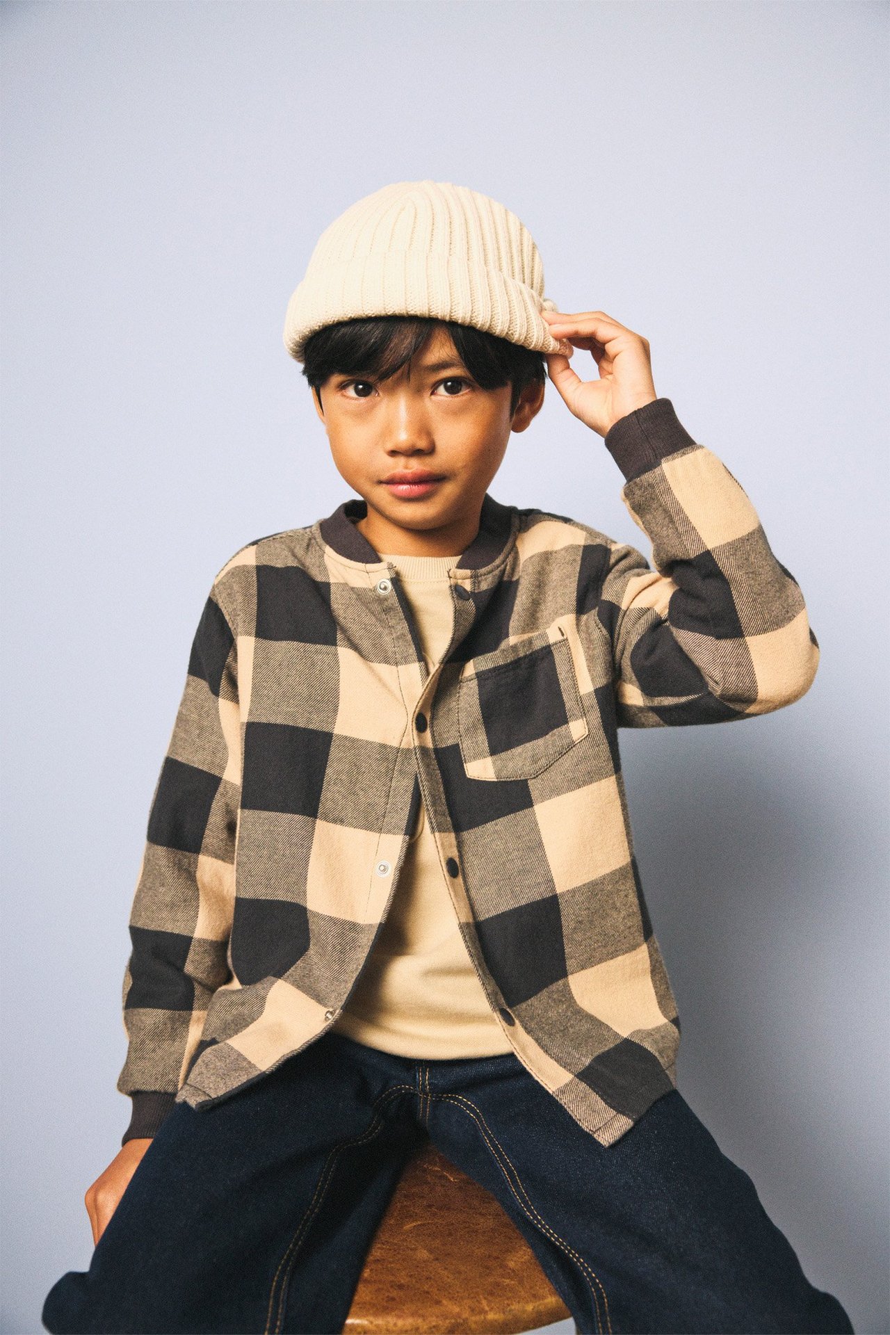 Kid med ljusbeige och mörkgrå rutig flanellskjorta, beige topp, mörka jeans och beige mössa.