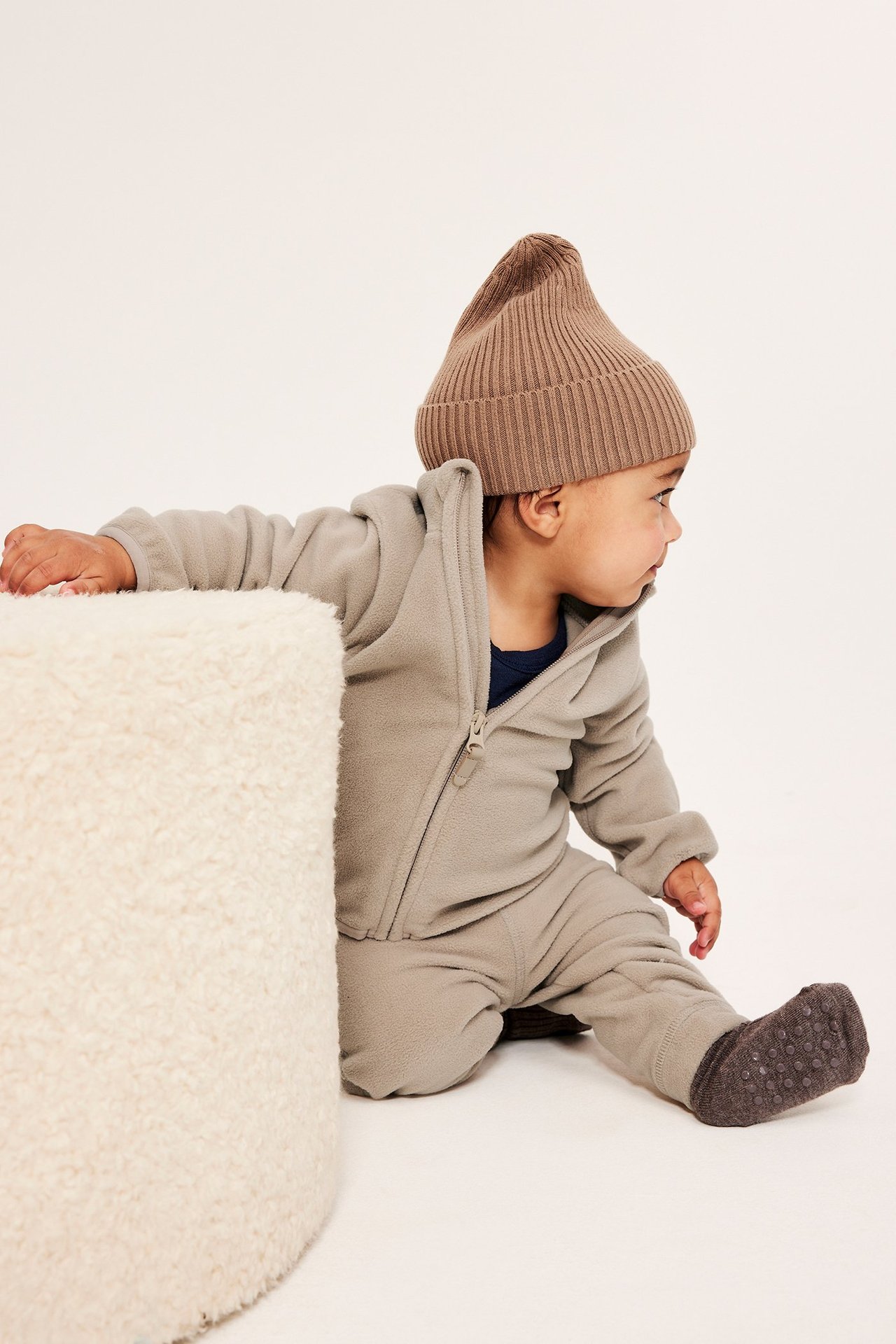 Sidevisning: Baby i beige fleece-heldress med hette, mørkeblå undertrøye, grå sokker, brun lue.