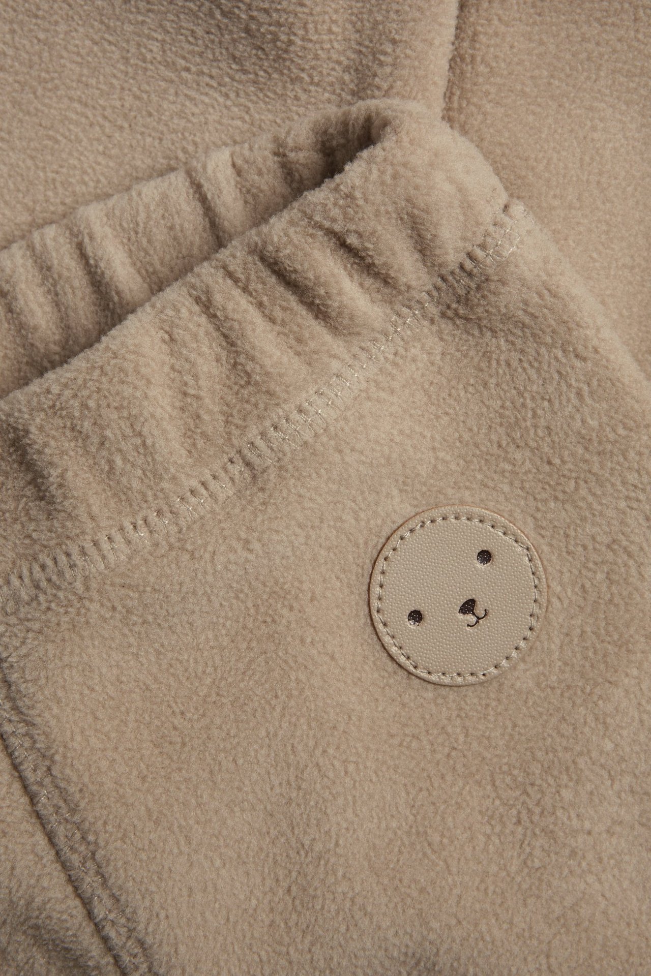 Zbliżenie beżowych spodni polarowych z elastyczną talią i małą okrągłą naszywką z misiem.