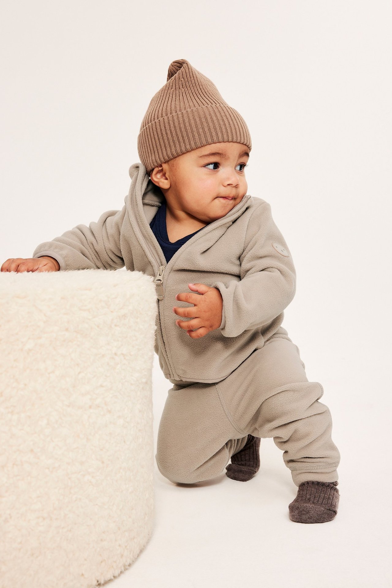 Forfra: Baby i beige fleecebukser, matchende hettejakke, brun ribbestrikket lue og mørkebrune tøfler.