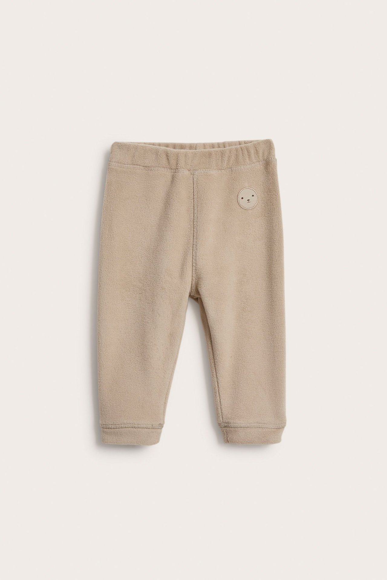 Frontvisning: Beige babyfleecebukse med elastisk midje og et lite rundt ansiktsmerke.