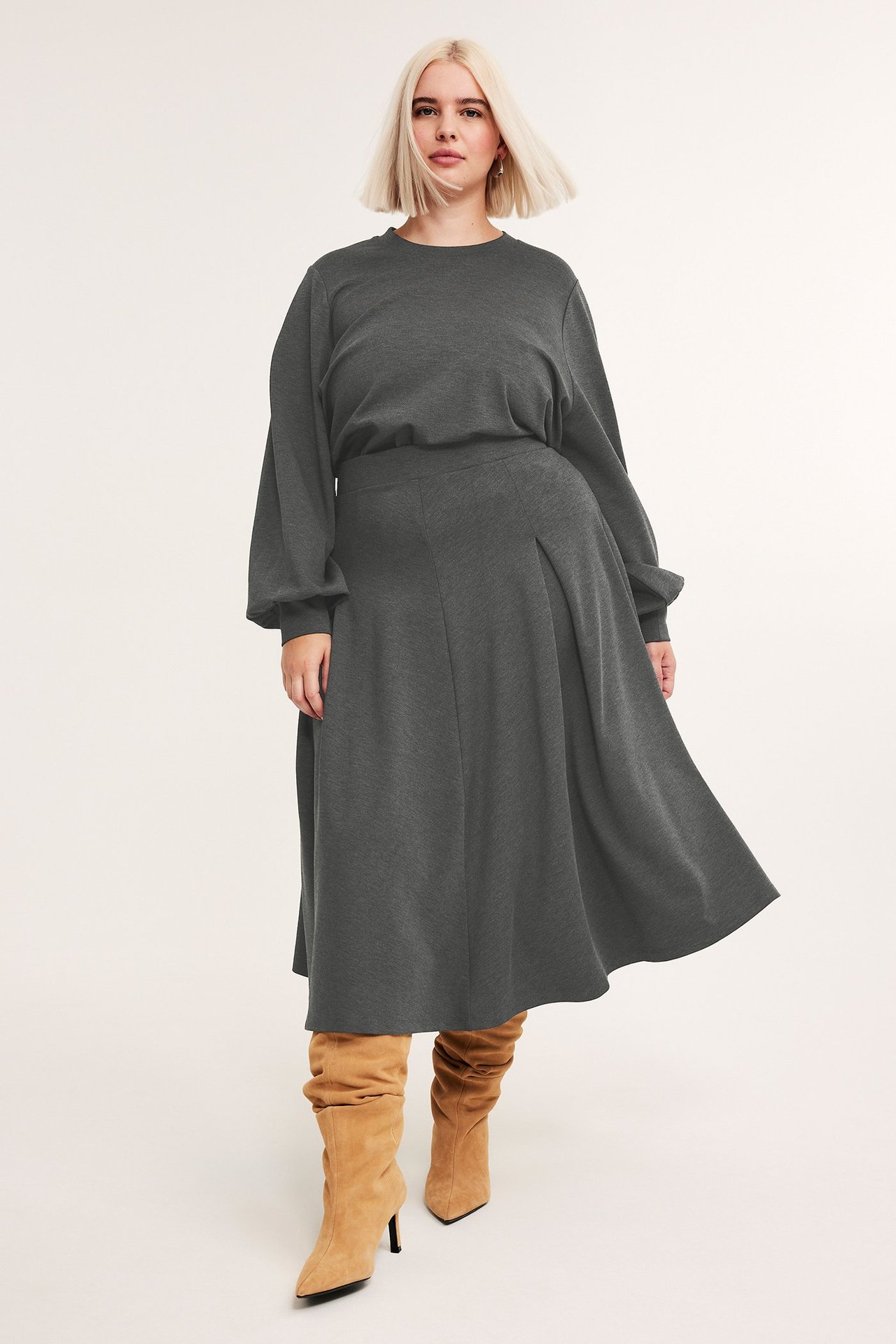 Edestä: nainen tummanharmaassa meleeratussa kellohameessa, yhteensopivassa pitkähihaisessa paidassa ja ruskeissa saappaissa.