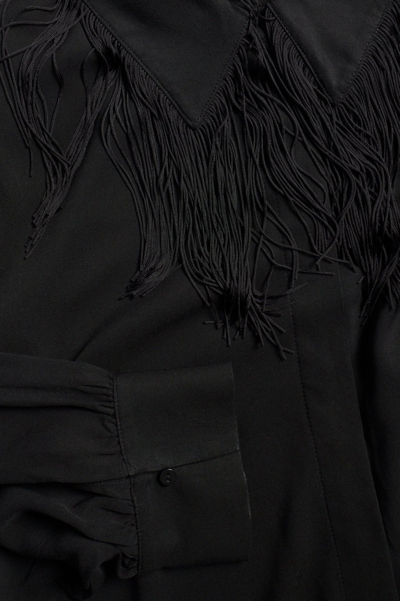 Zbliżenie czarnej krótkiej koszuli z frędzlami przy kołnierzu i długimi rękawami z mankietami na guziki.