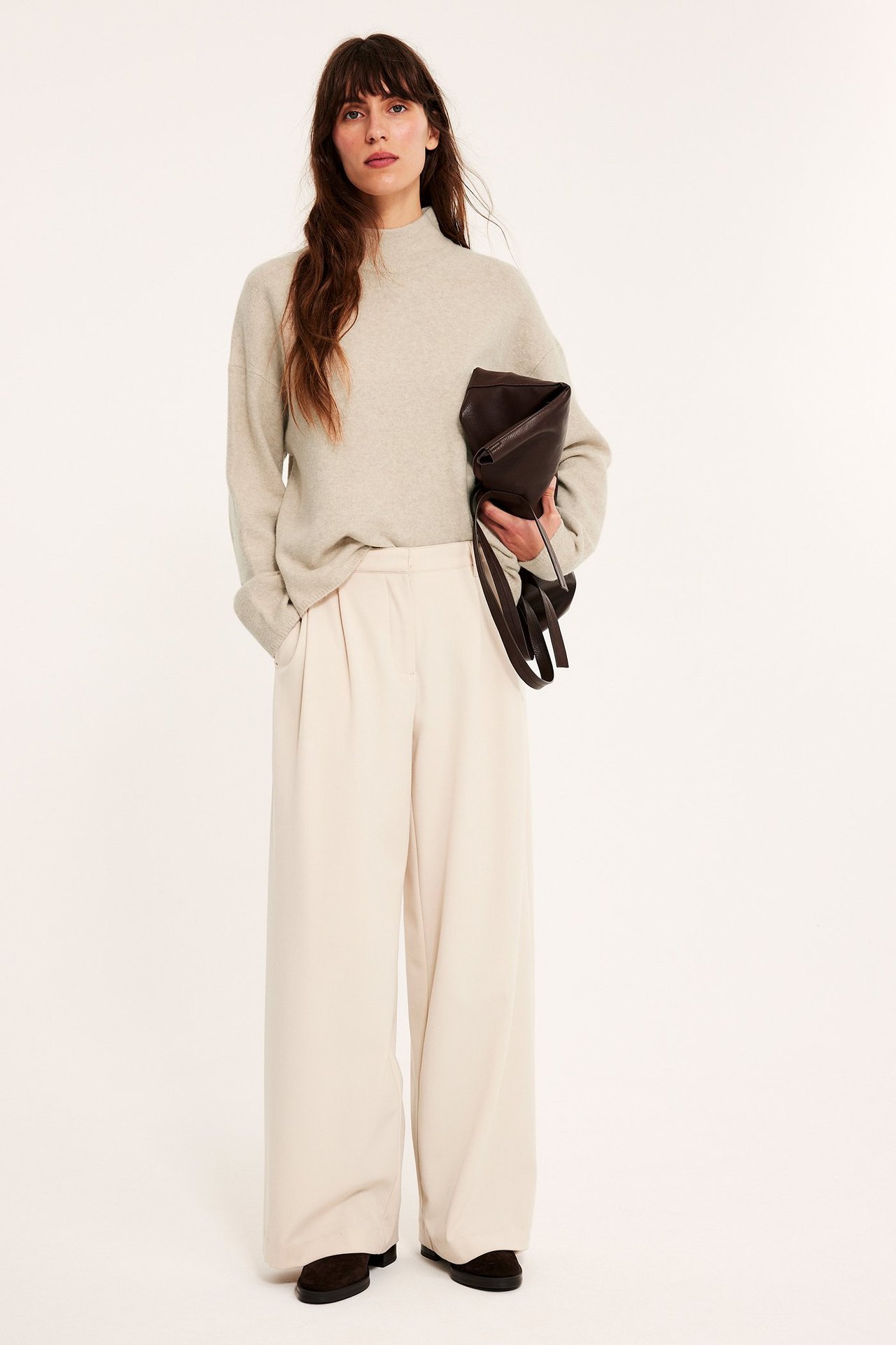Forfra: Kvinne i beige melert strukturert turtleneck-genser, kremfargede vide bukser, holder mørk clutch.