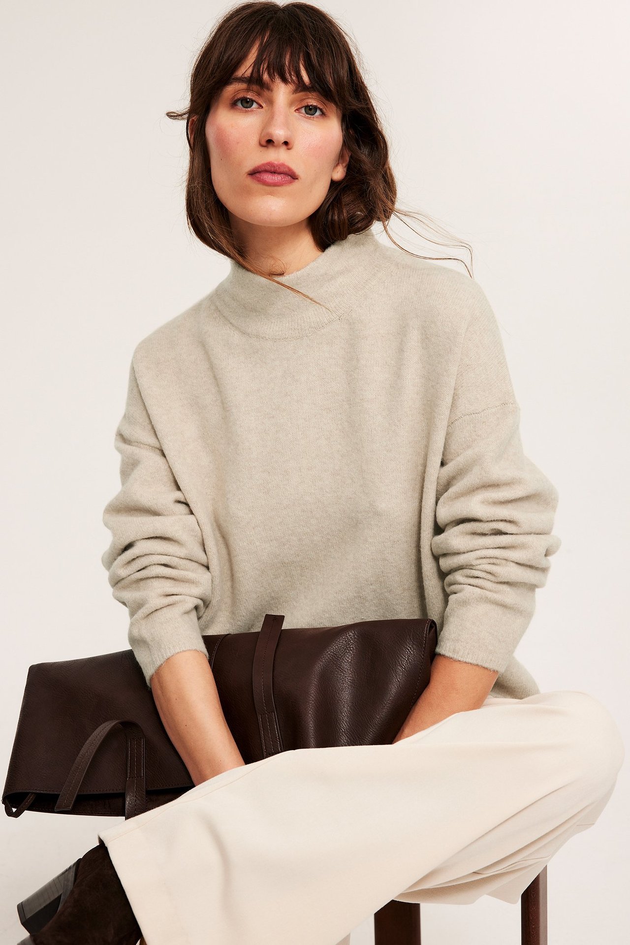 Forfra: kvinne i melert beige strukturert turtleneck-genser, lyse bukser, holder en brun veske.