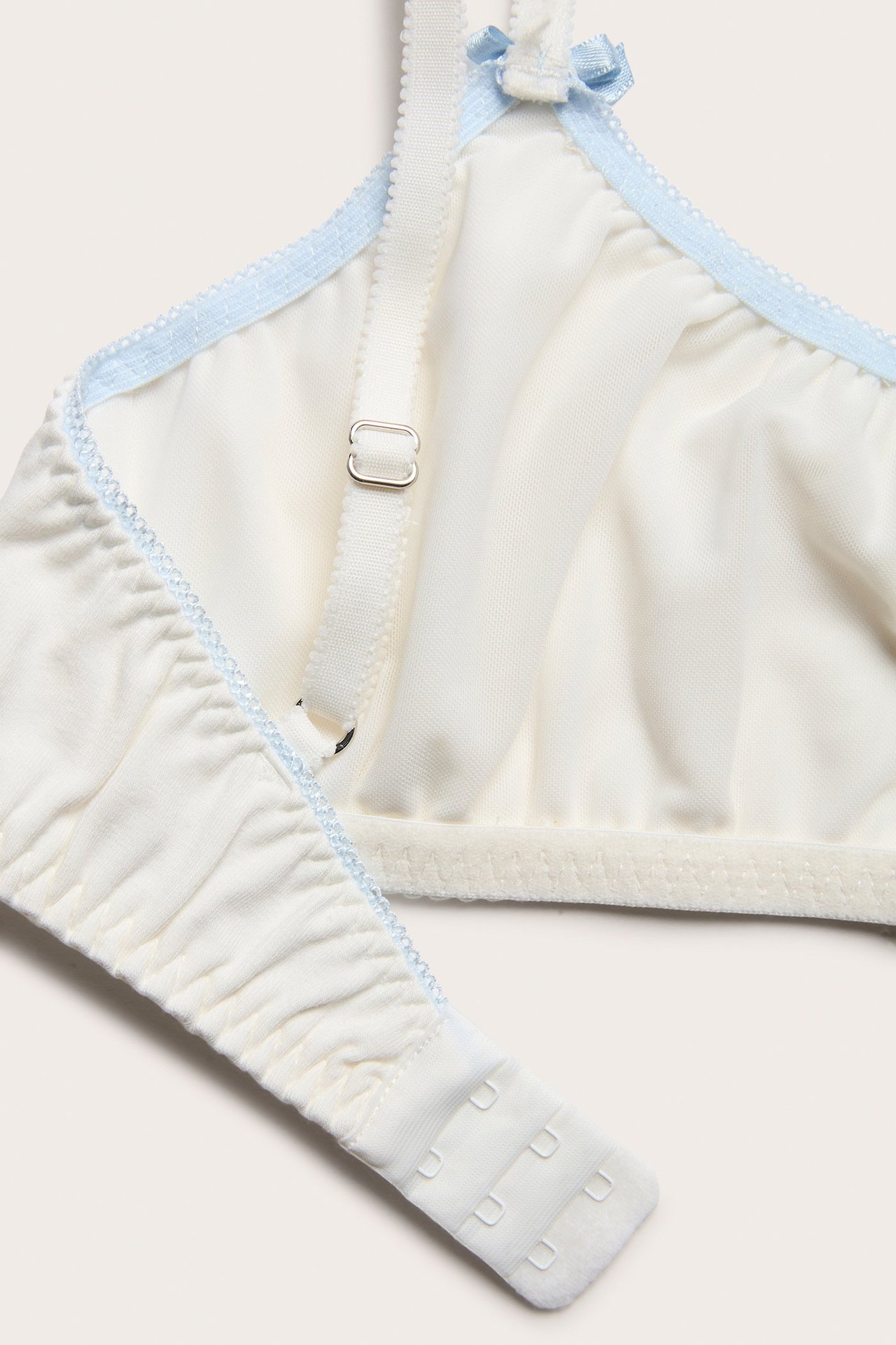 Zbliżenie białego miękkiego biustonosza z jasnoniebieskim obszyciem, regulowanymi ramiączkami, małymi kokardkami i zapięciem z tyłu.