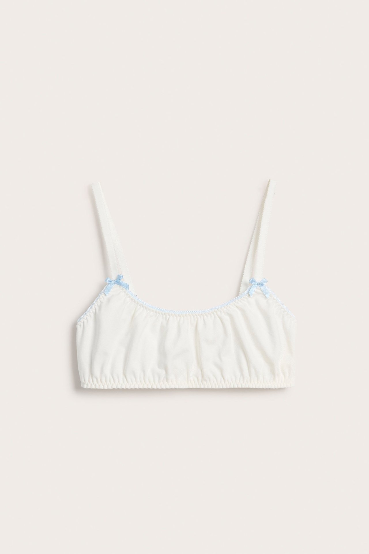 Framifrån: vit mjuk bralette med rynkad tyg, ljusblå kantband och små blå rosetter.