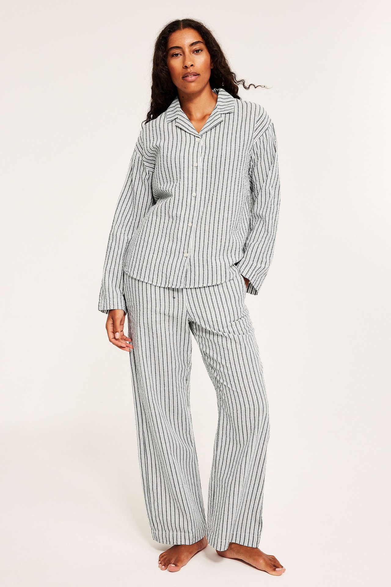 Edestä näkymä naisesta yllään valko- ja tummaraidallinen puuvillapyjamassetti pitkähihaisella paidalla ja housuilla.