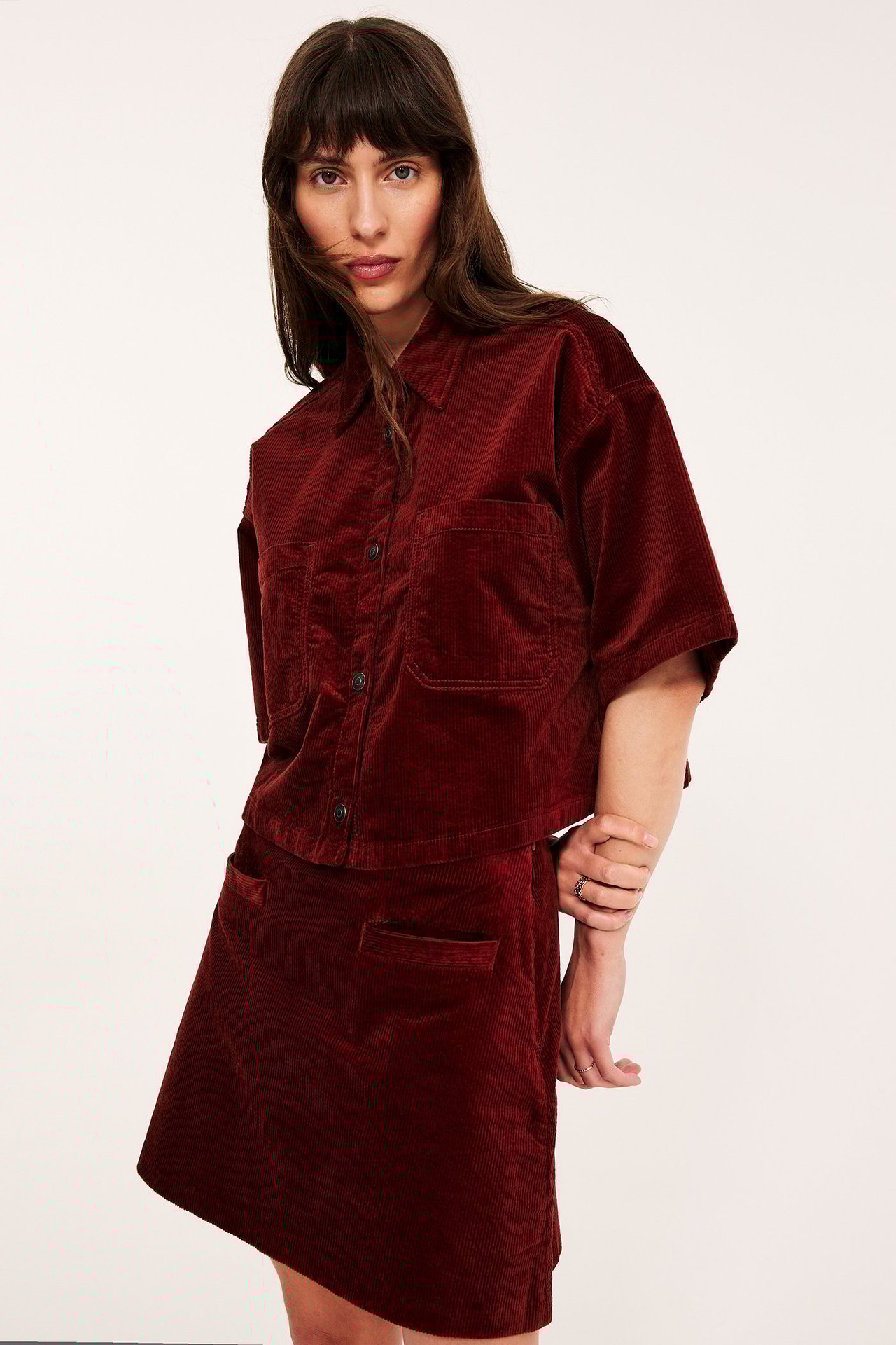 Frontbilde av kvinne i mørkerødt kordfløyel miniskjørt med lommer og matchende kortermet skjorte.