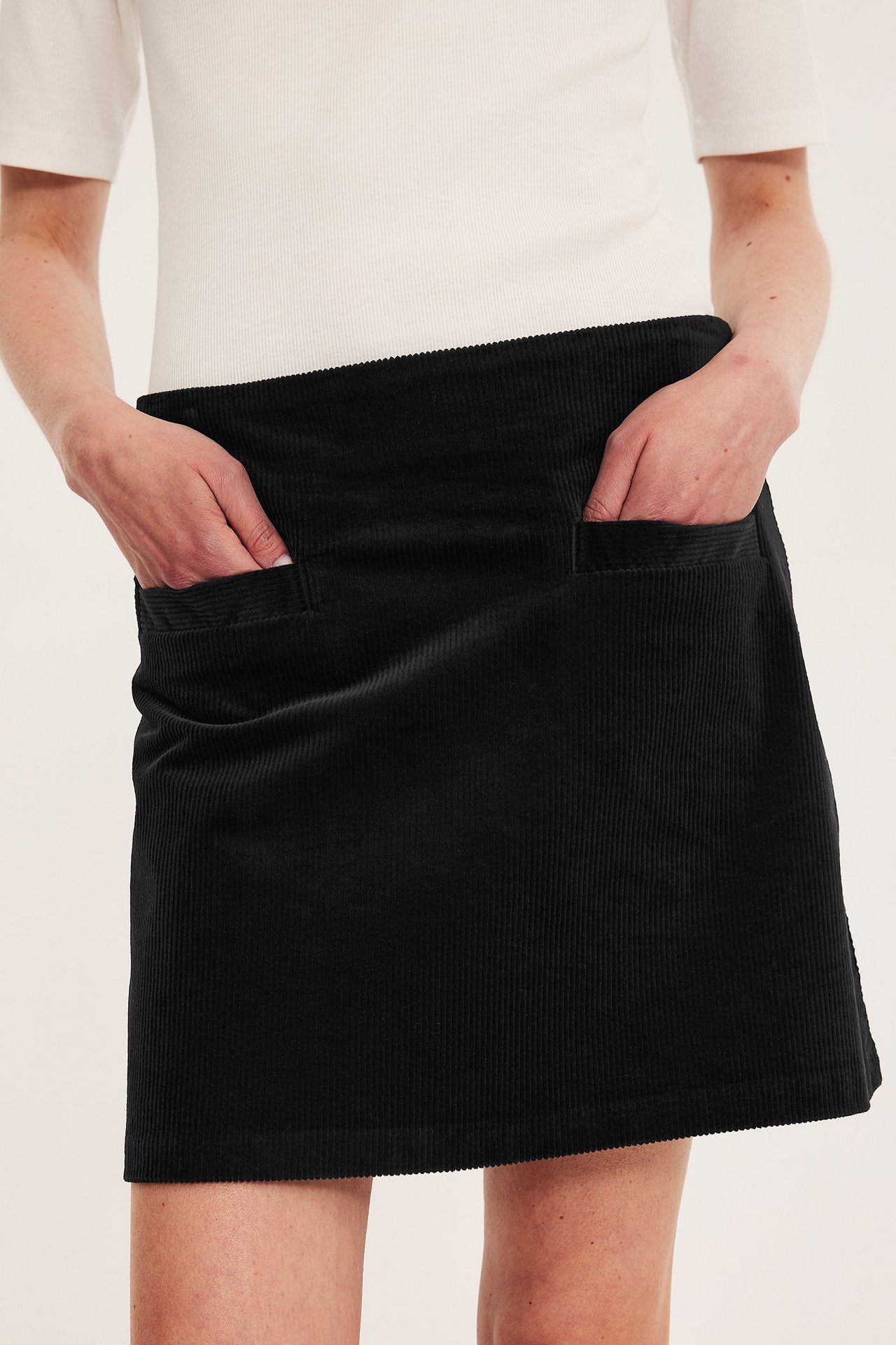 Frontbilde av en kvinne i mørkeblått kordfløyel miniskjørt med frontlommer og en hvit ribbet topp.