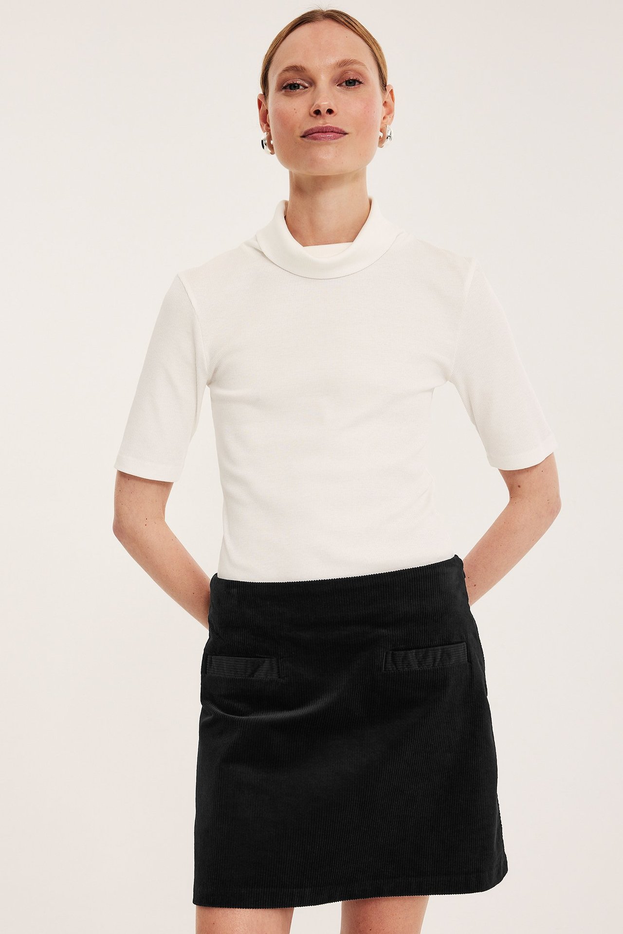 Frontbilde: Kvinne i mørkeblå kordfløyel miniskjørt med frontlommer og hvit kortermet topp.