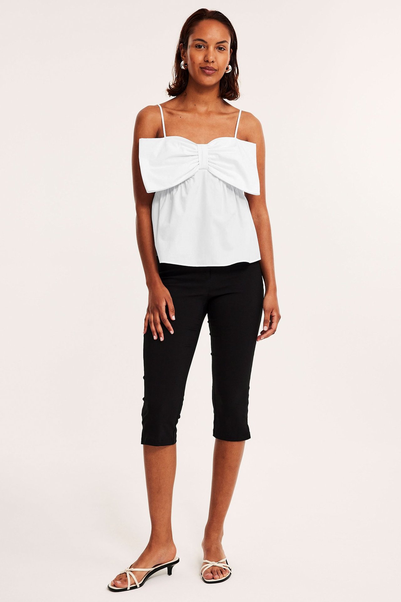 Forfra: kvinne i hvit peplumtopp med stor sløyfe, svarte capribukser og hvite sandaler.