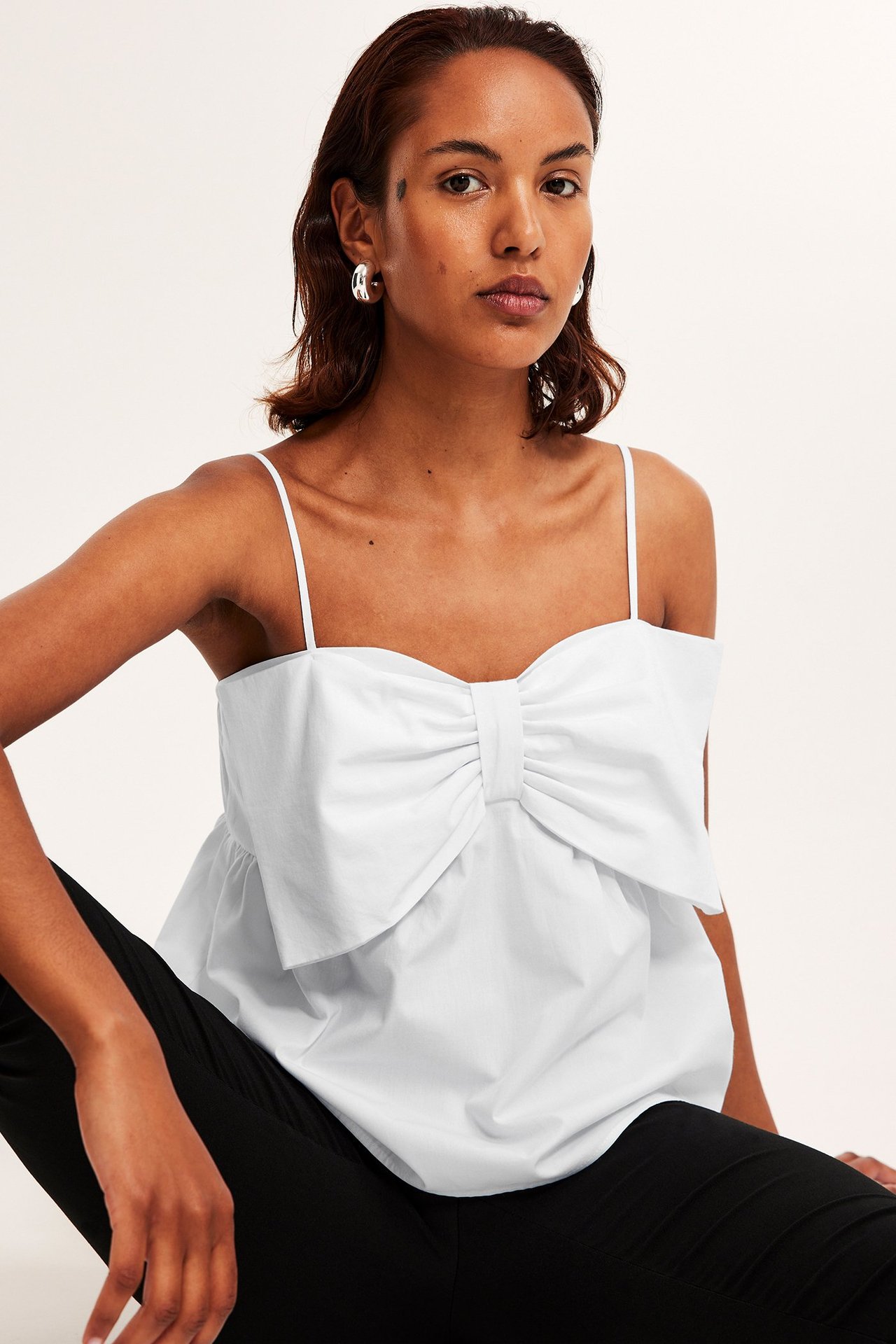 Forfra: kvinne i hvit peplumtopp med stor sløyfe, svarte bukser og sølvfargede ringøreringer.