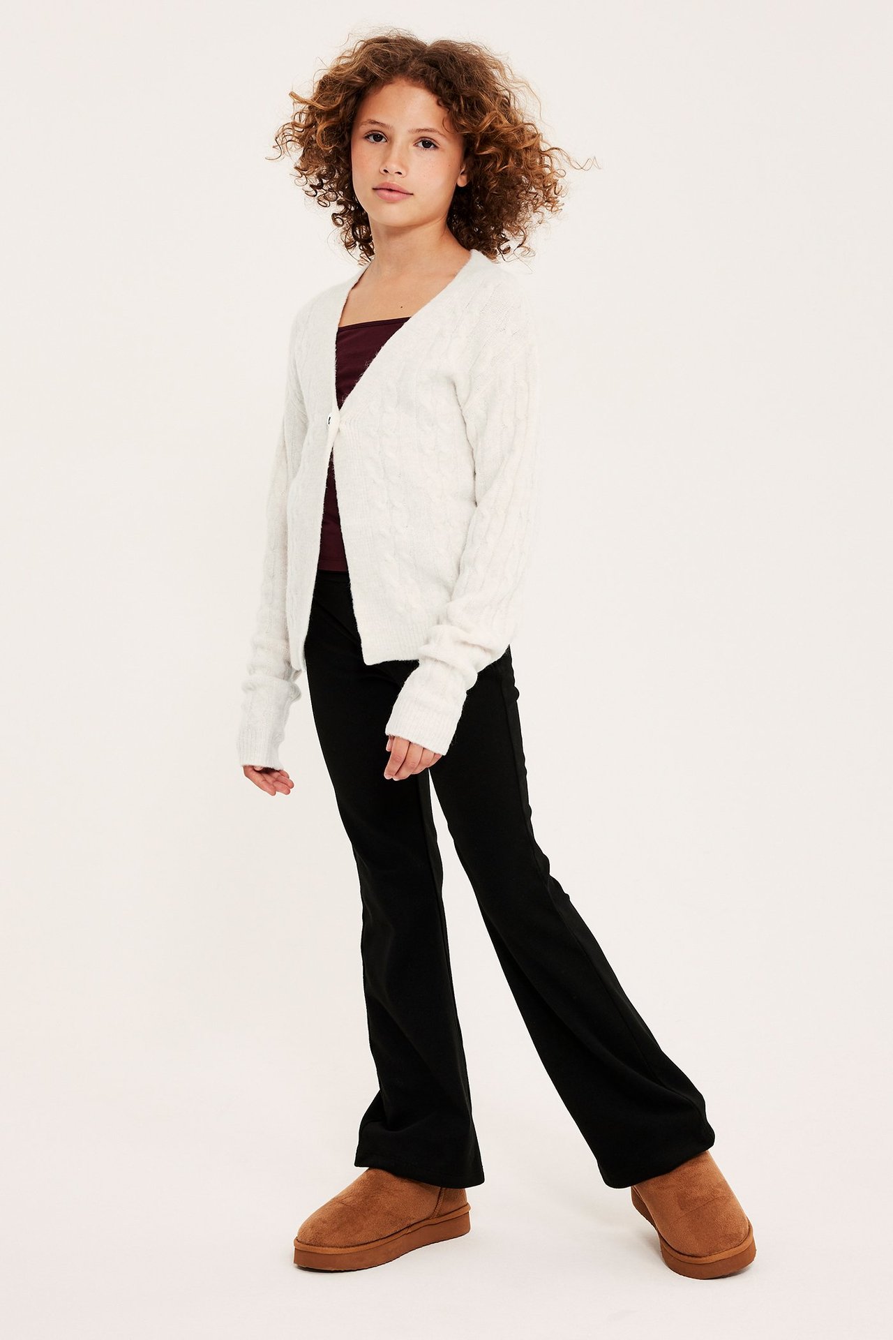 Barn i vit kabelstickad kofta med hjärtknapp, mörkröd topp, svarta byxor och bruna stövlar.