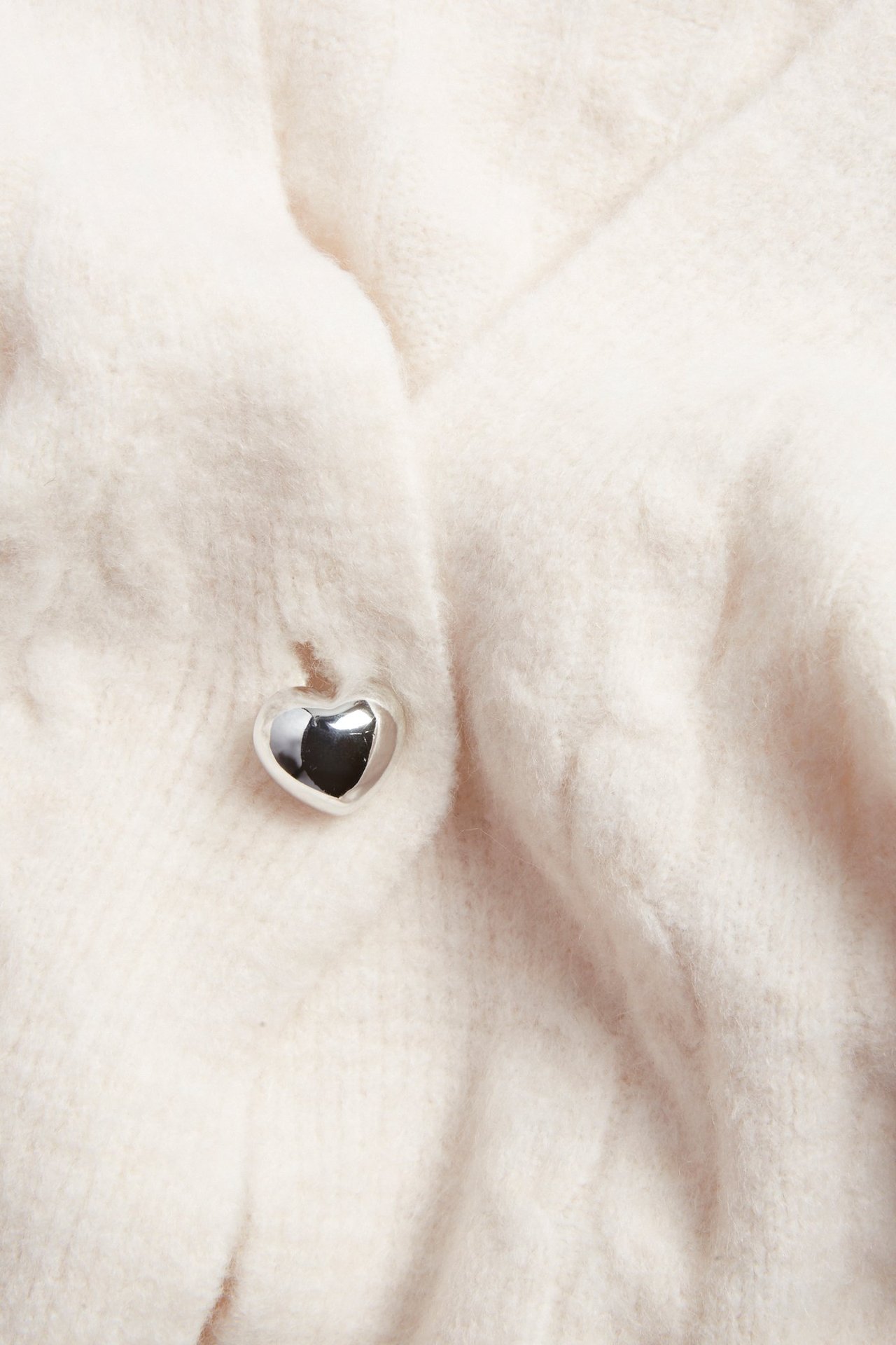 Zbliżenie miękkiego, białego, melanżowego kardiganu w warkocze z srebrnym guzikiem w kształcie serca.