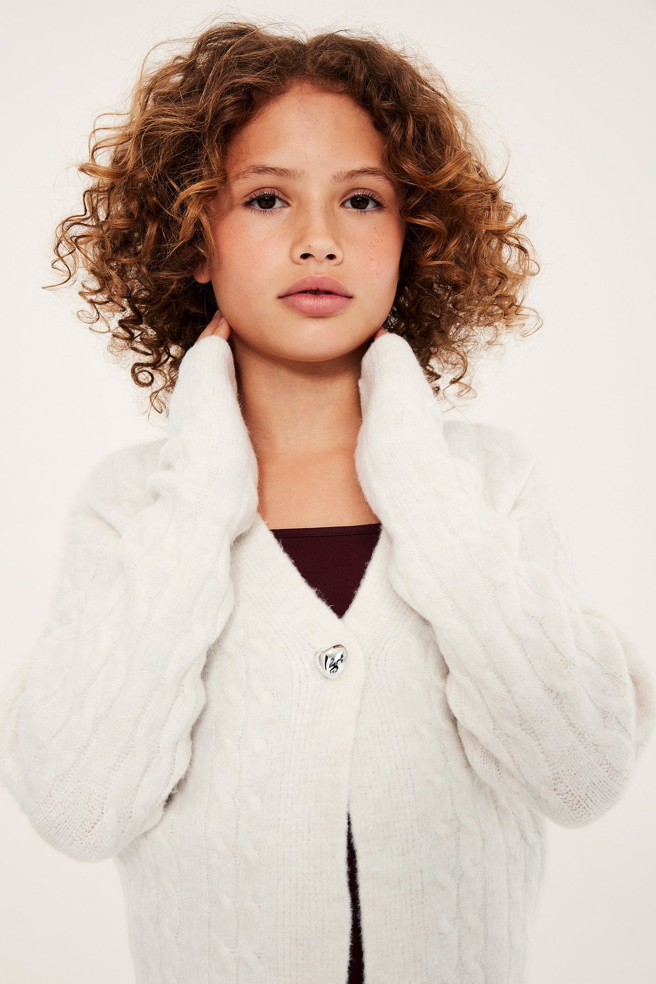 Närbild framifrån på ett barn i vit kabelstickad kofta med hjärtknapp över en mörkröd topp.