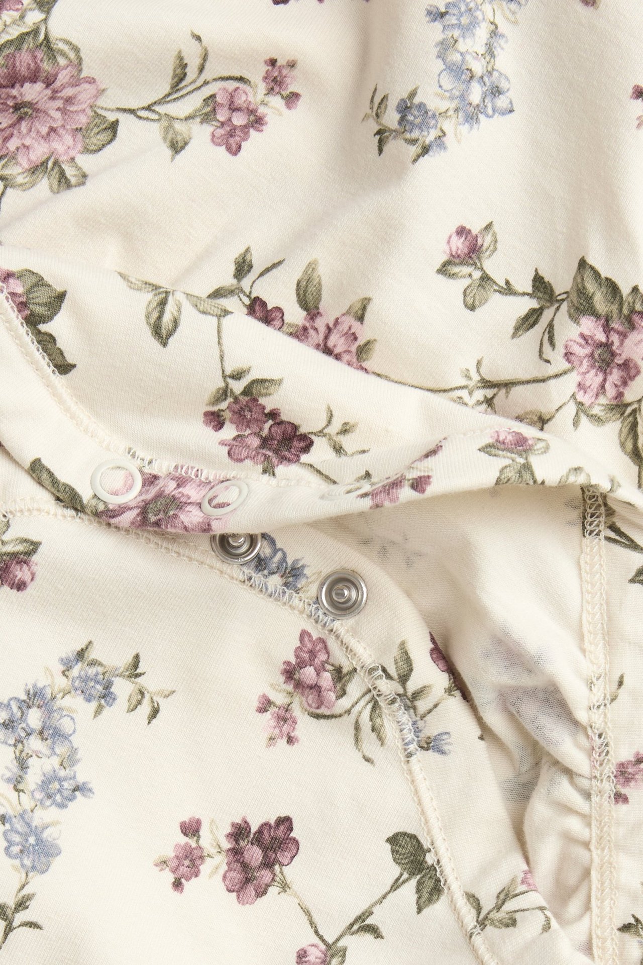 Zbliżenie na jasnobeżowe body niemowlęce z fioletowo-niebieskim wzorem kwiatowym i zatrzaskami.