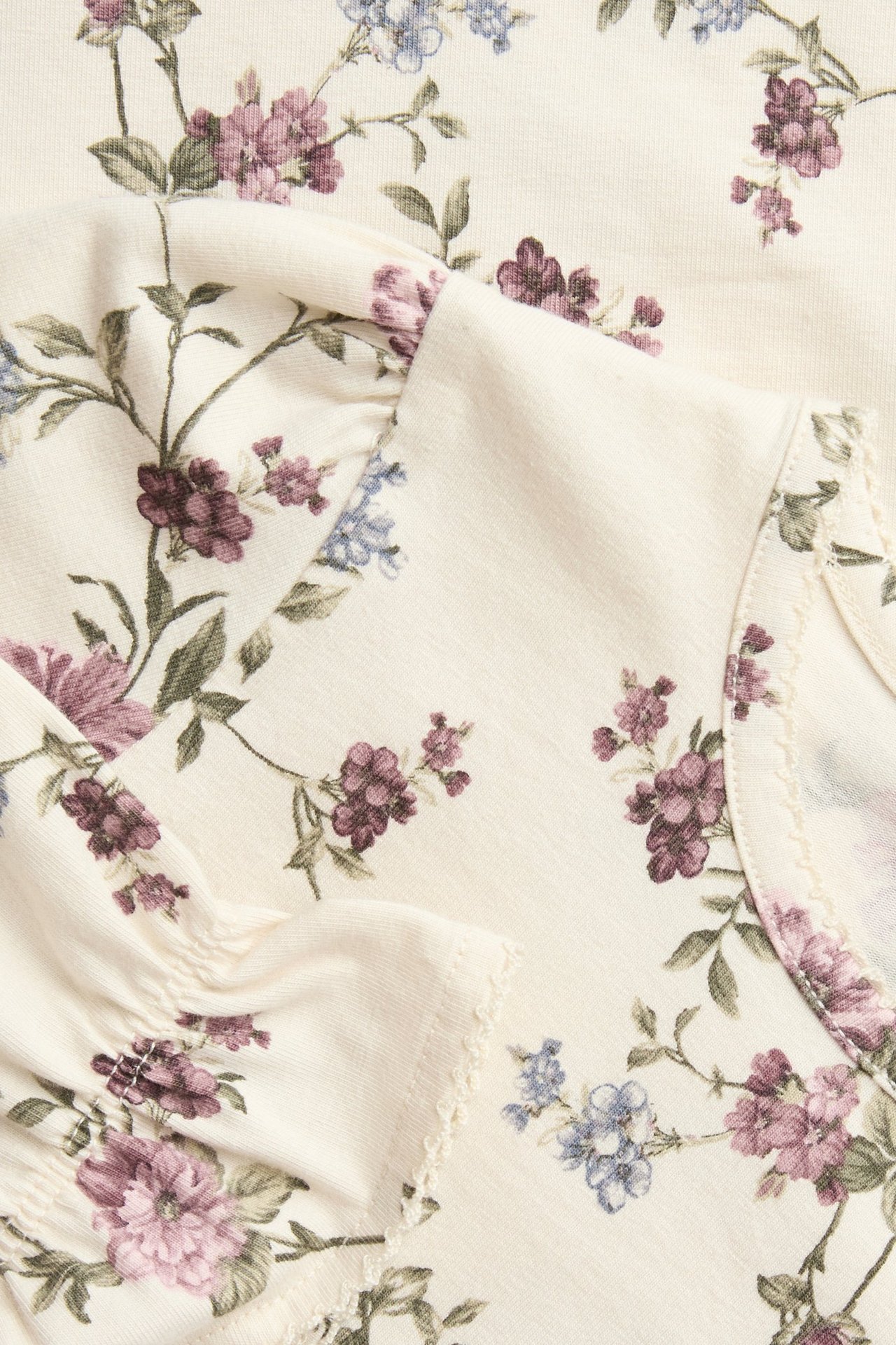 Nærbilde av en lys beige blomstret topp med lilla og blå blomster, puffermer og picotkanter.