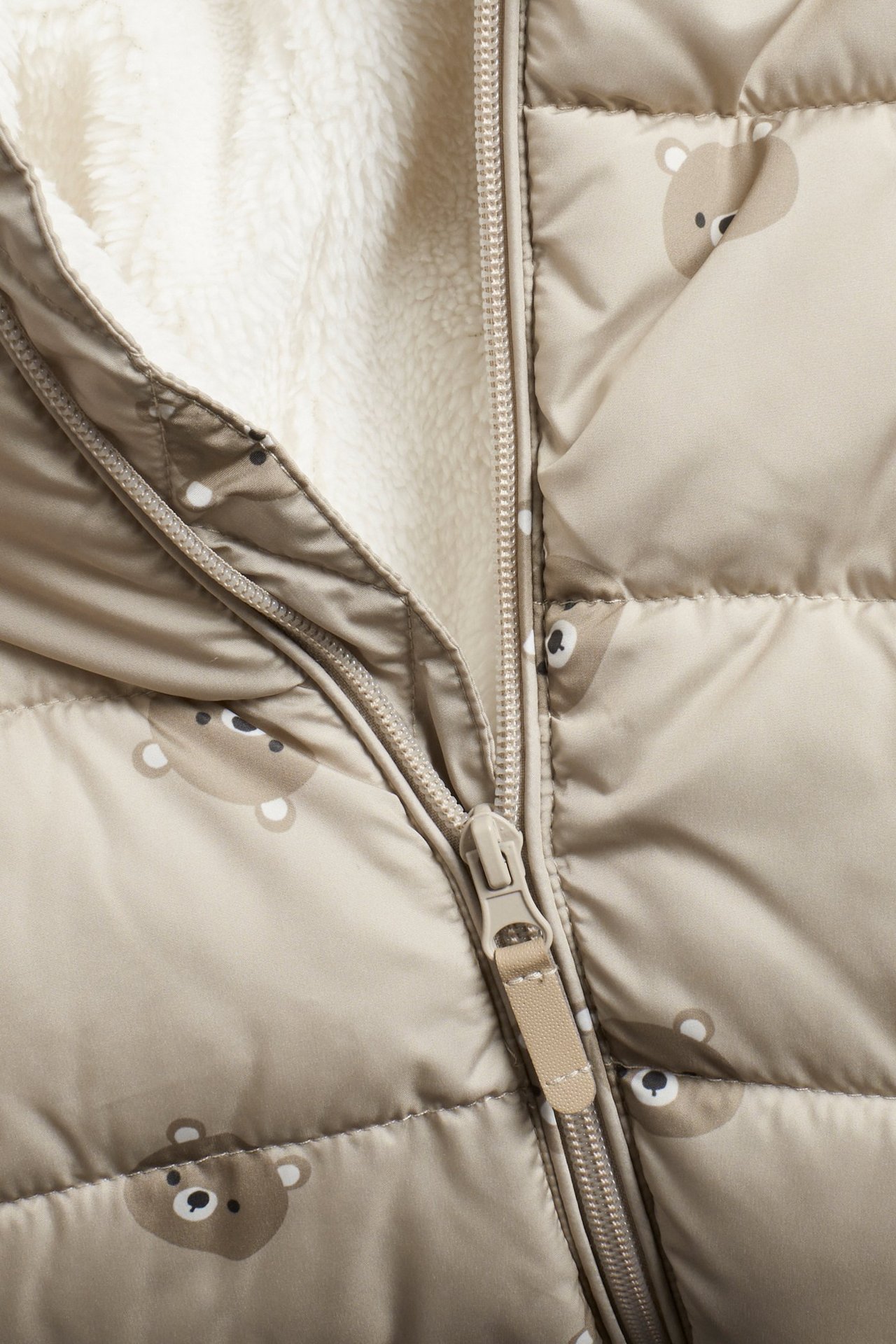 Närbild på beige vadderad babyoverall med björnmönster och synligt krämfärgat pilefoder.