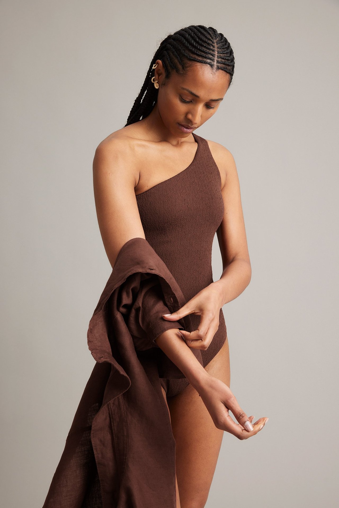 Framifrån: Kvinna i mörkröd enaxlad baddräkt med struktur, håller matchande skjorta, med guldfärgade örhängen.