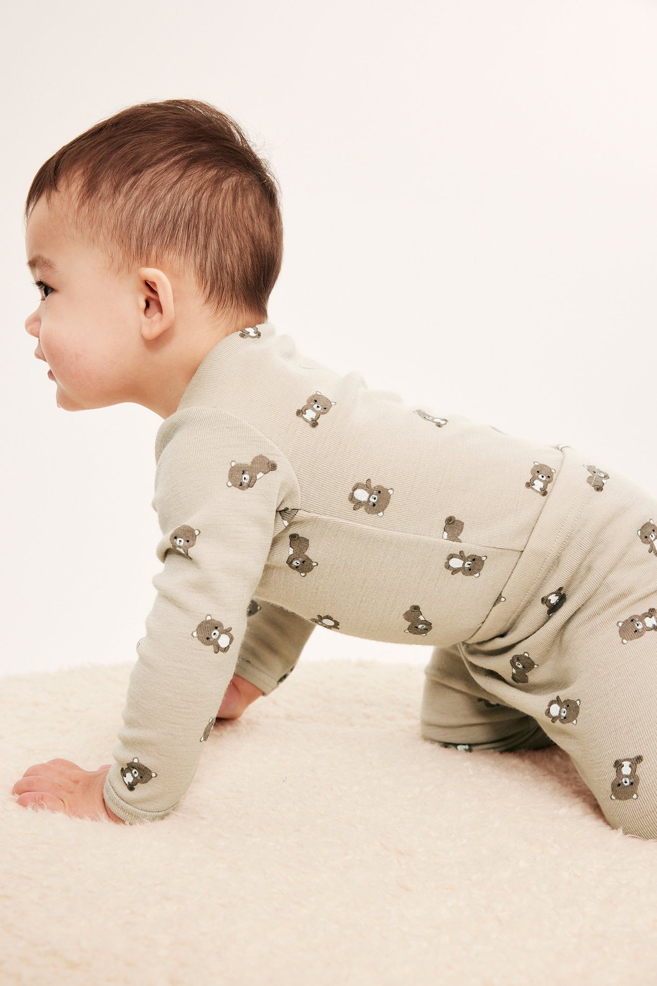 Sidebilde av en krabbende baby i en beige merinoullsbody og matchende bukser med bjørnemønster.